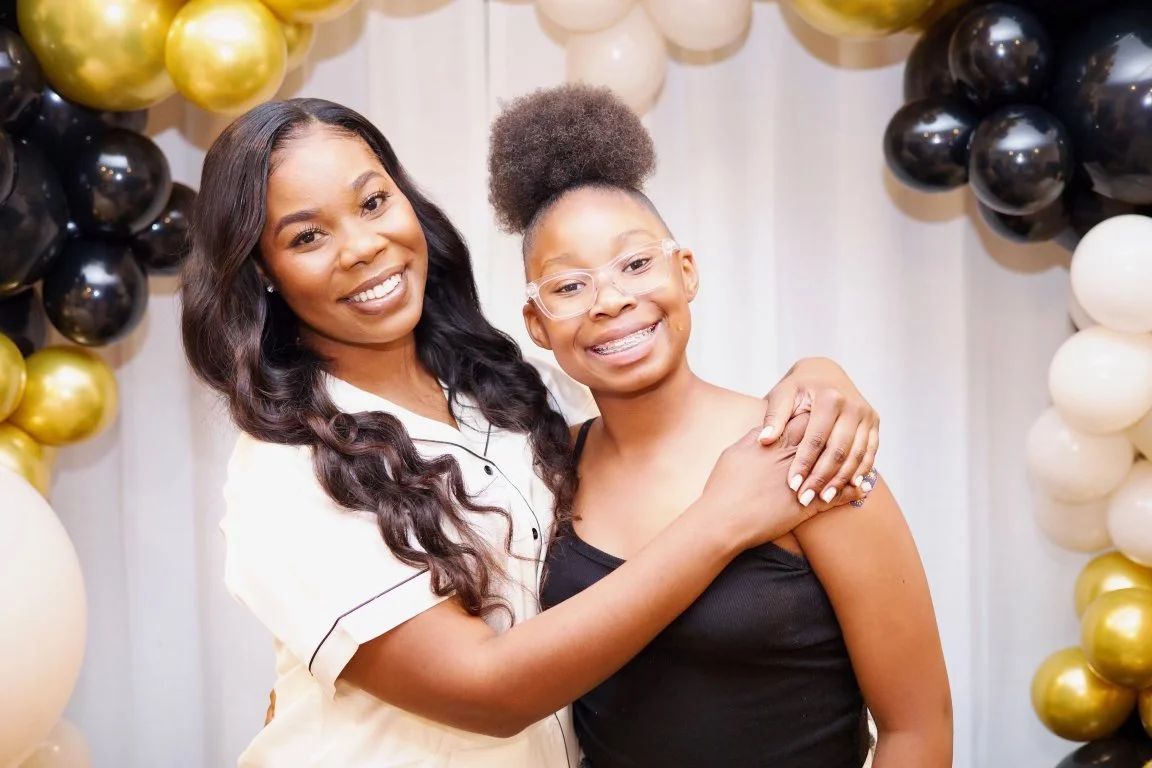 Two women smiling and embracing in front of a balloon backdrop with black, white, and gold balloons.