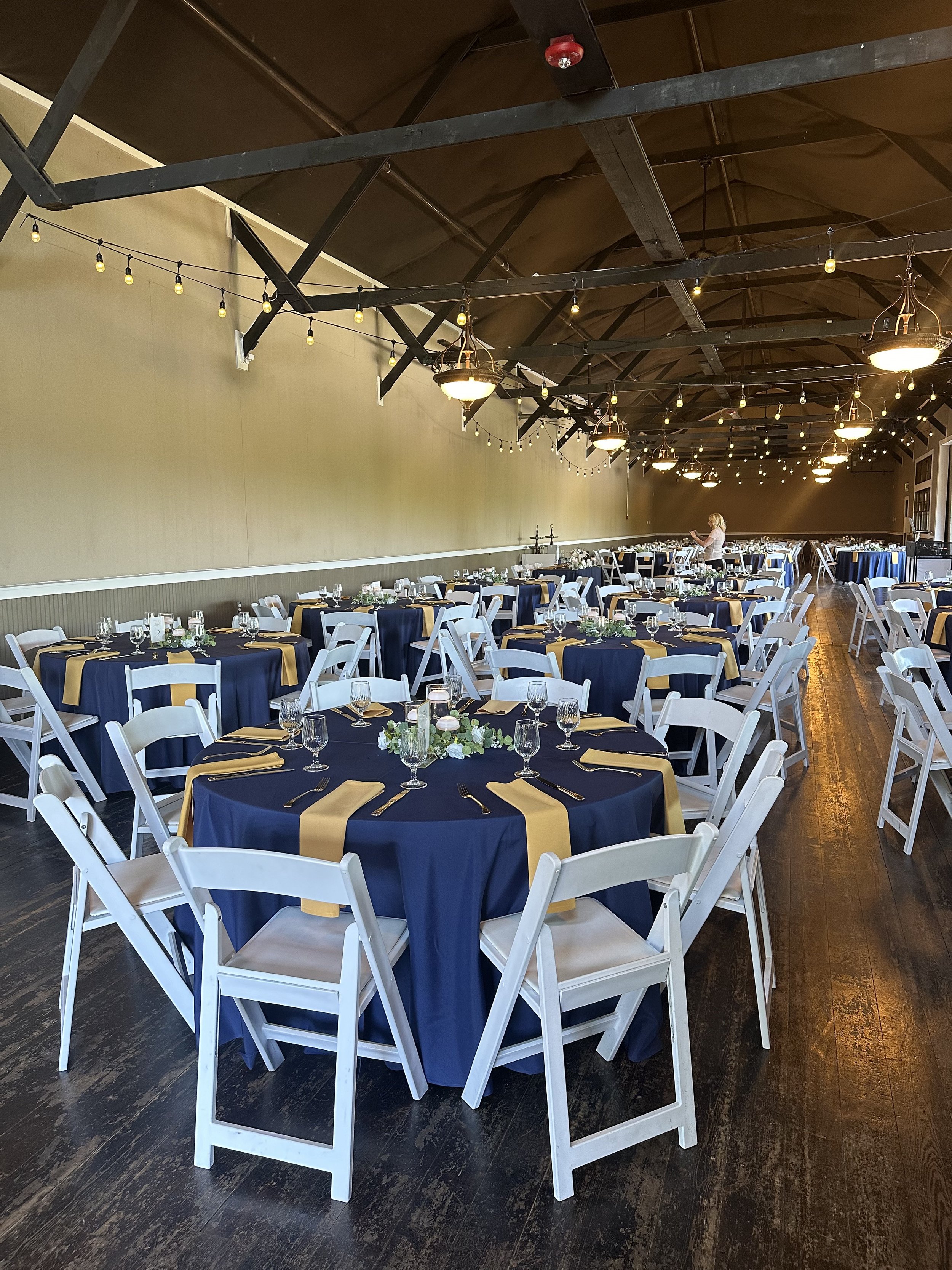 Elegant event hall decorated with round tables covered in navy blue tablecloths and gold runners, white chairs, floral centerpieces, and string lights hanging from the ceiling.