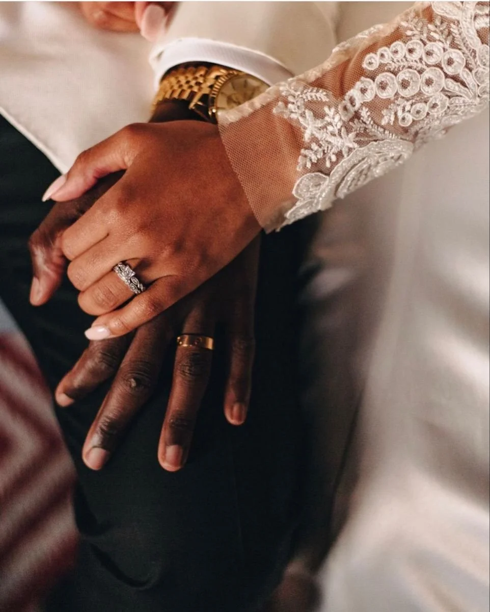 Close-up of a couple holding hands, showcasing wedding rings, with the woman wearing a lace sleeve and a gold watch, and the man dressed in a white shirt and dark suit.