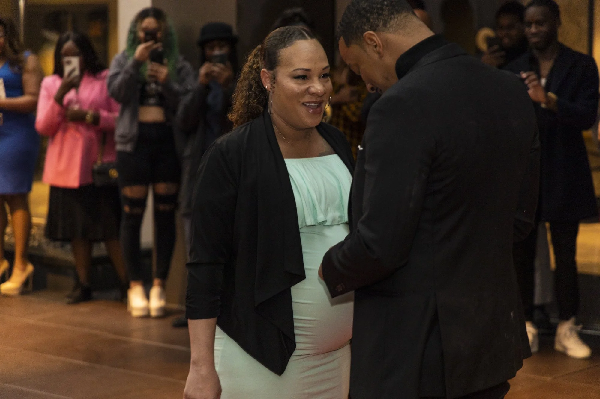 A woman and a man sharing a special moment at a social gathering, with people taking photos in the background.