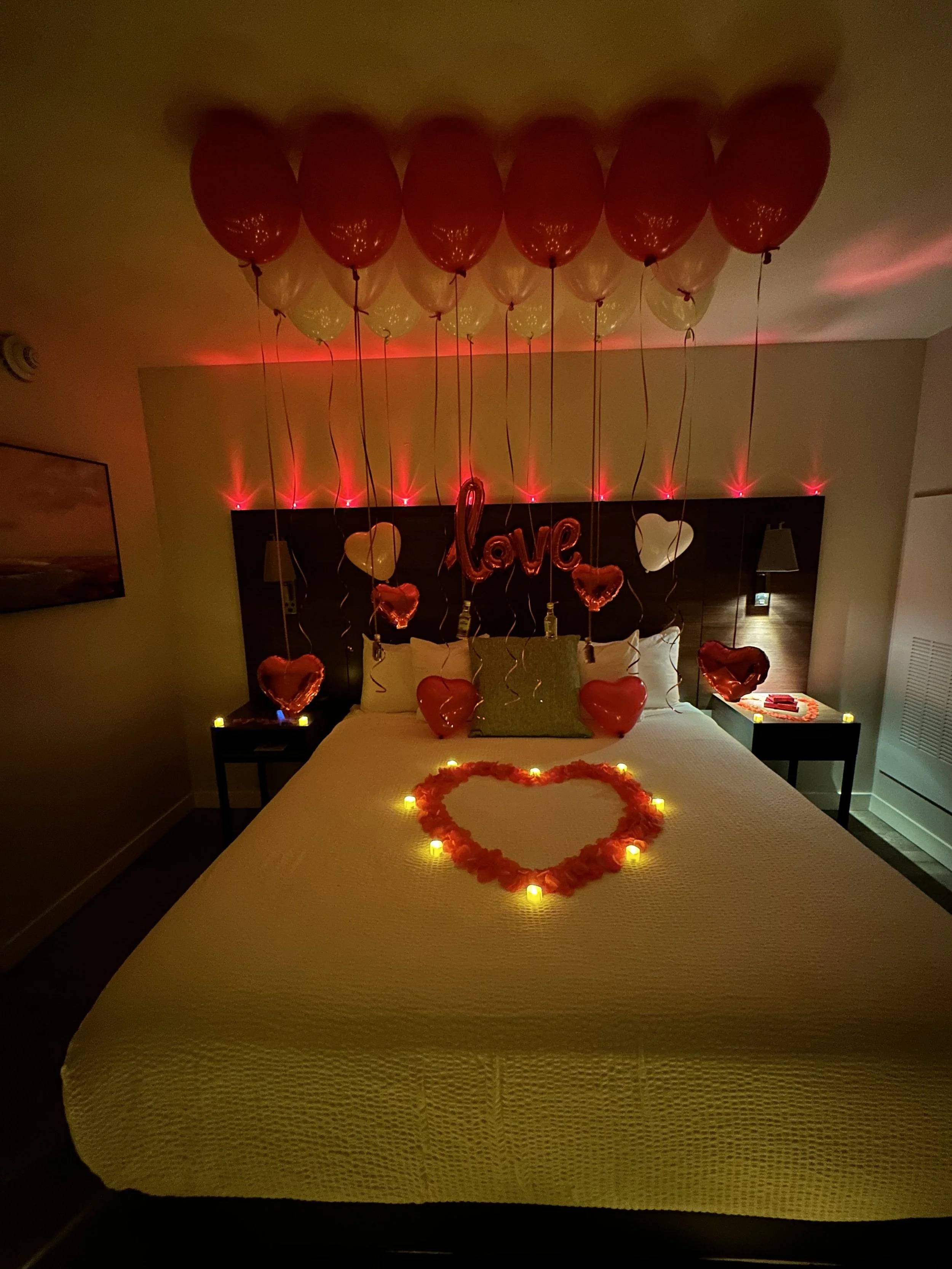 Romantic hotel room decorated with red and white balloons, heart-shaped pillows, LED candles forming a heart on the bed, and a "Love" balloon ornament hanging from the ceiling.