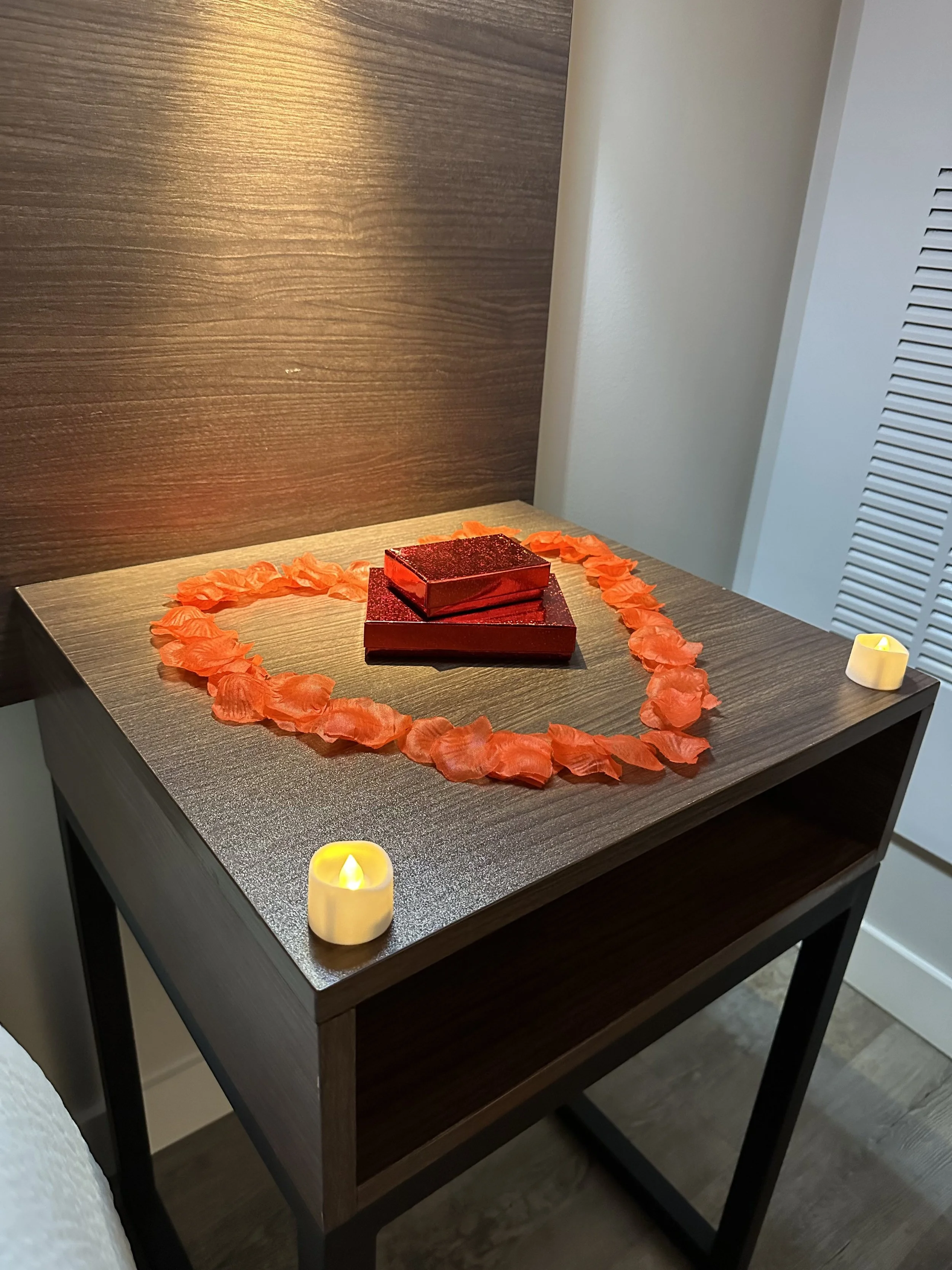 Decorative table display with red-themed items, surrounded by orange rose petals arranged in a heart shape, and two small candle lights on each corner.