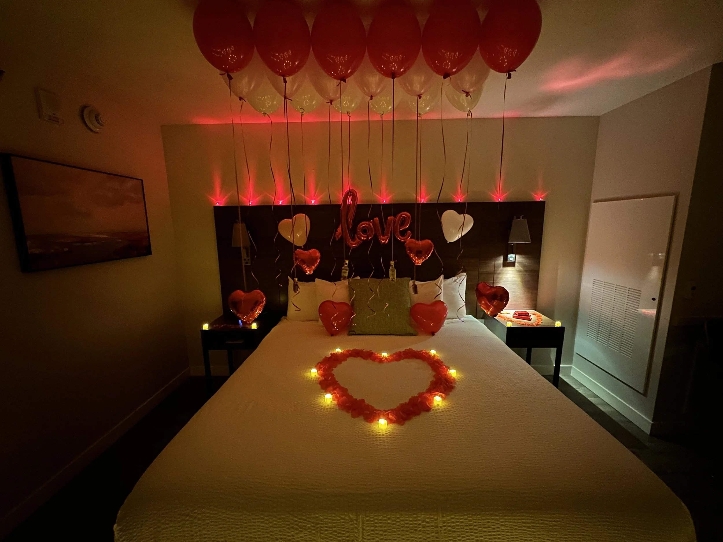A hotel room decorated with balloons and lights for Valentine's Day, featuring heart-shaped balloons, a 'love' balloon, candles, and LED lights arranged in a heart shape on the bed, with candles and small items on the nightstands.