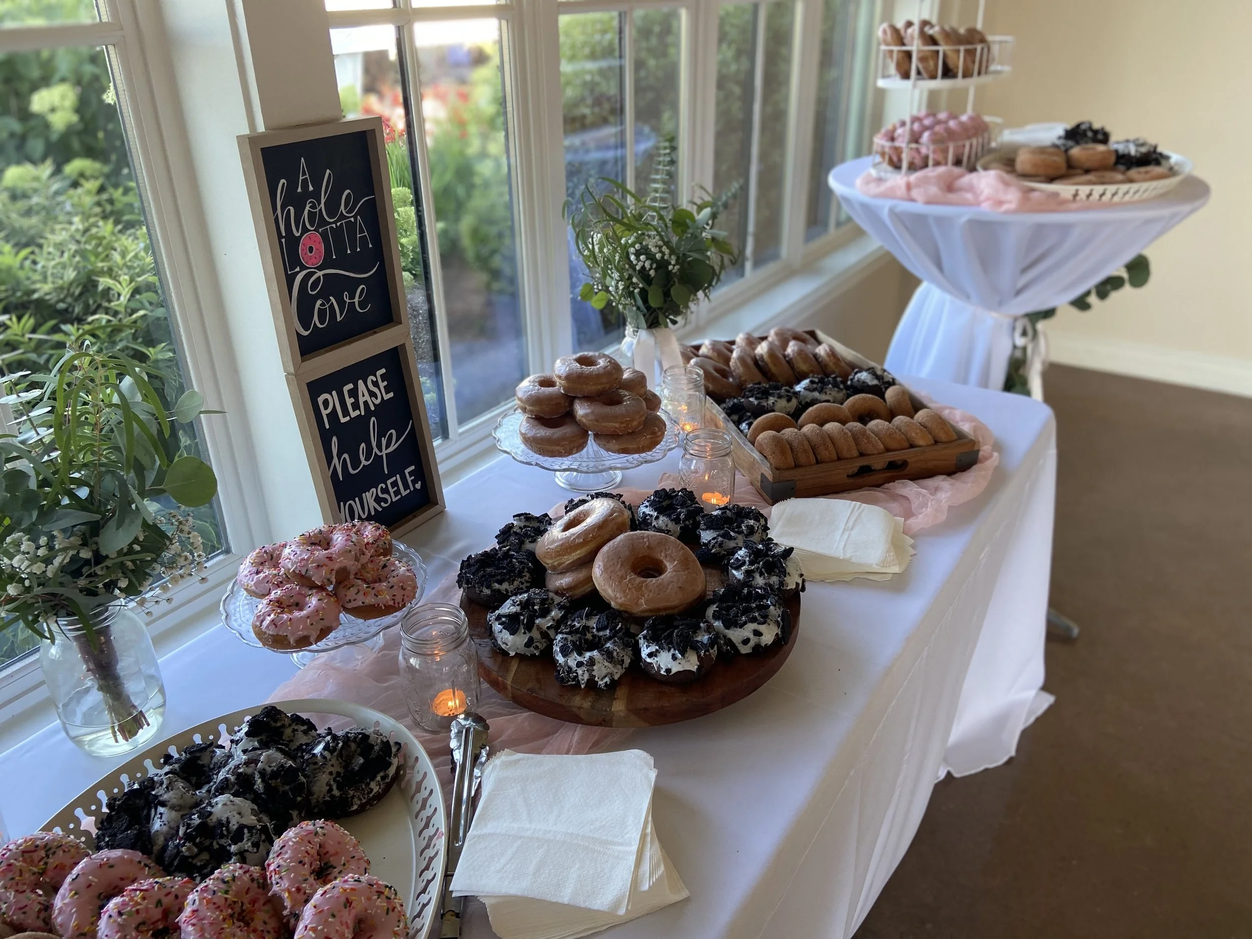 A table set with various donuts and baked goods, decorated with a pink and white theme, near windows with greenery outside, and a sign that reads 'A hole lotta love, please help yourself.'
