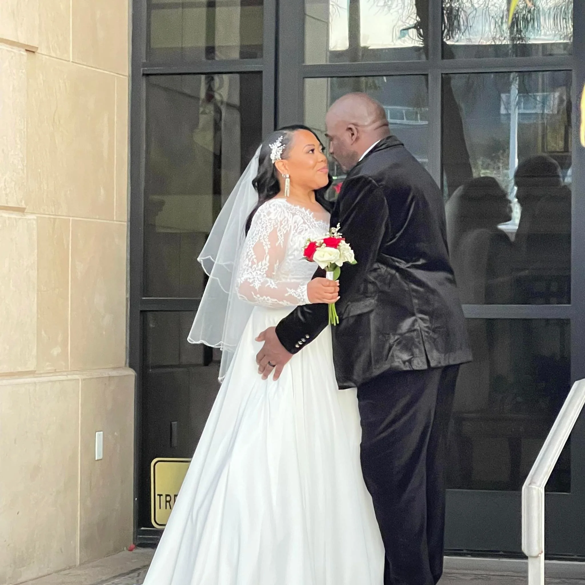A bride and groom embrace outside a building, gazing into each other's eyes. The bride wears a white lace wedding dress with long sleeves and a veil, holding a small bouquet of red and white flowers. The groom wears a black suit and jacket, standing 