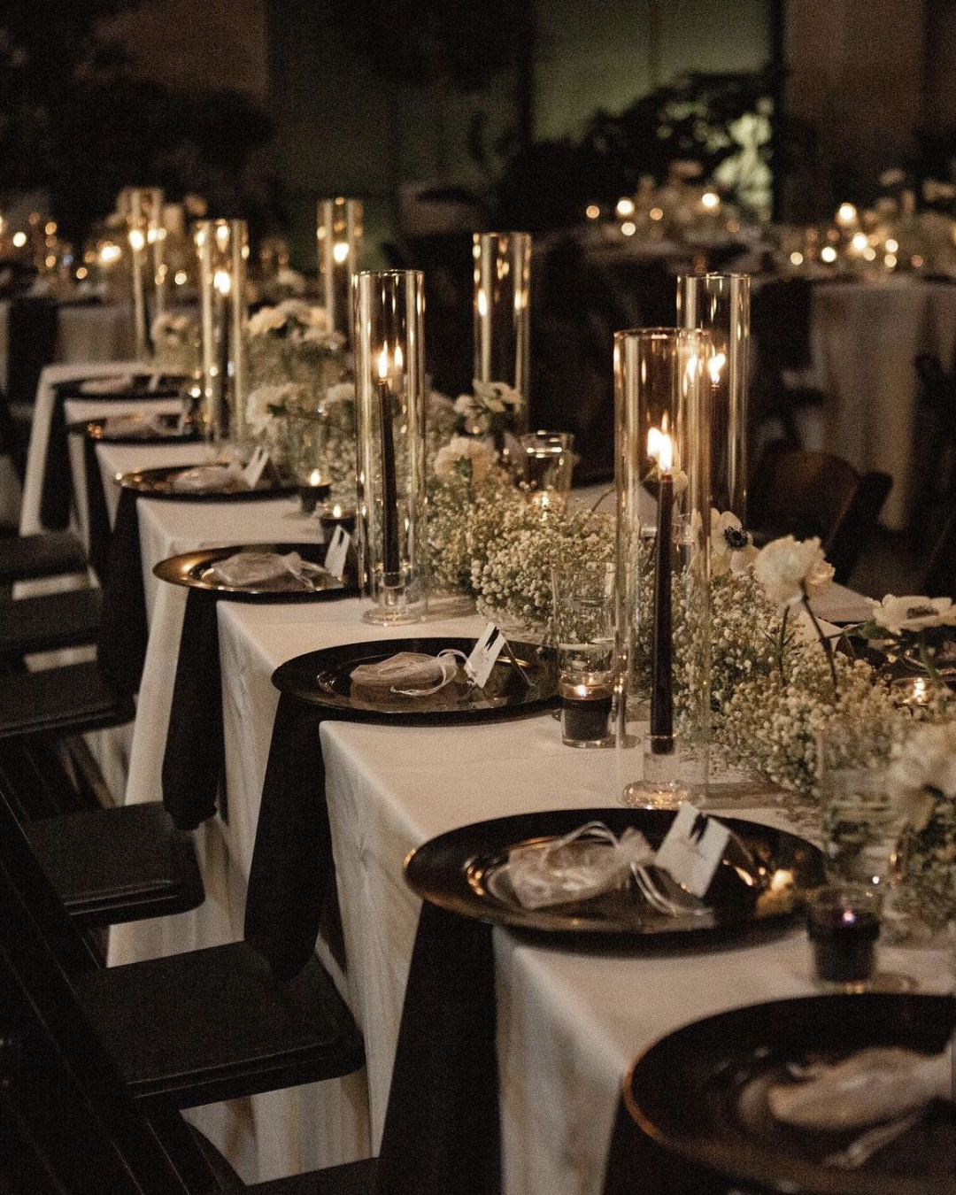 Elegant banquet table with black plates, white napkins, gold utensils, tall candles, and floral centerpieces in a dimly lit setting.