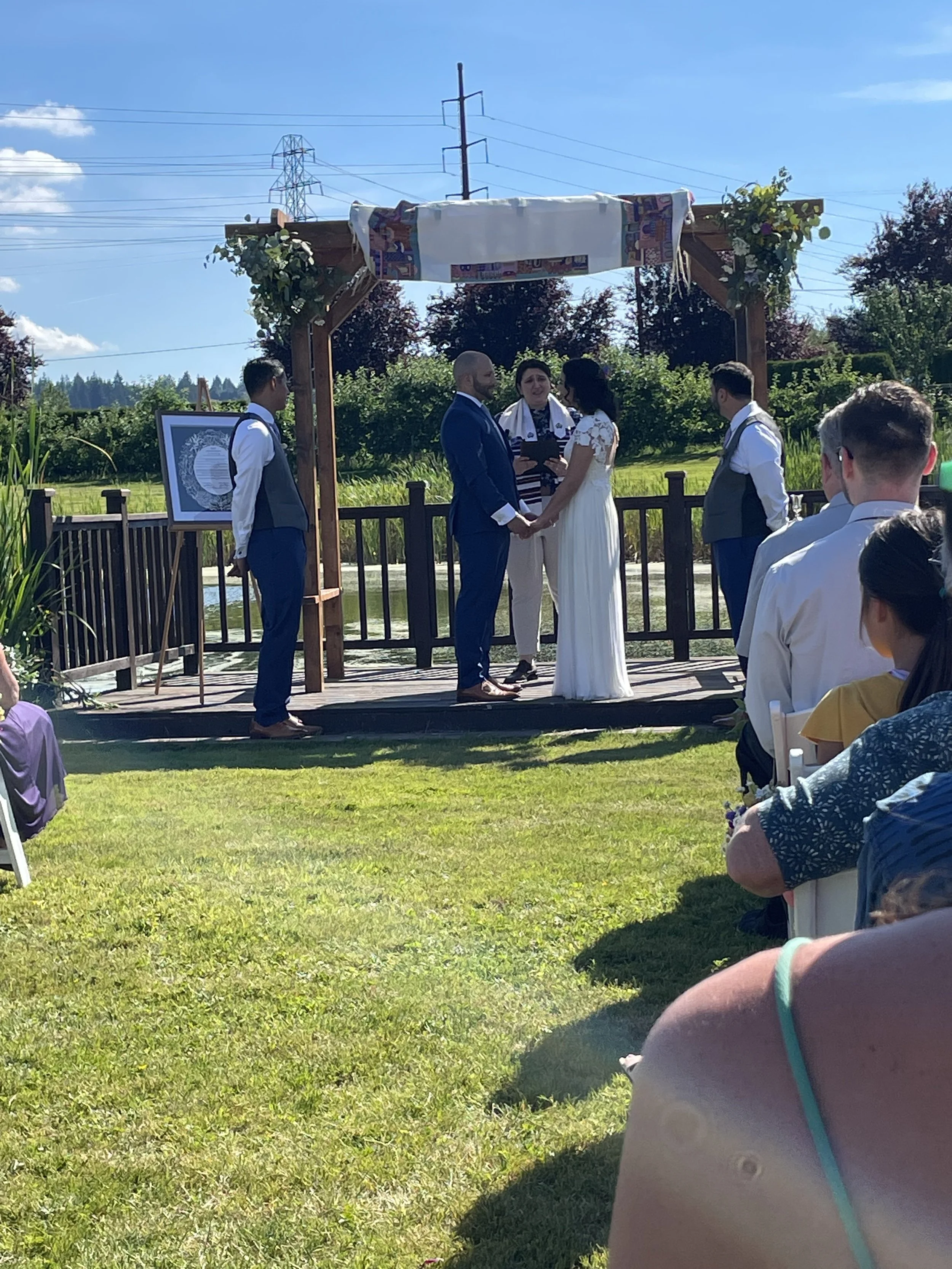 A couple gets married outdoors, standing underneath a wooden arch decorated with greenery and fabric, holding hands and facing each other while an officiant conducts the ceremony. Guests are seated on the grass, watching the event on a sunny day with