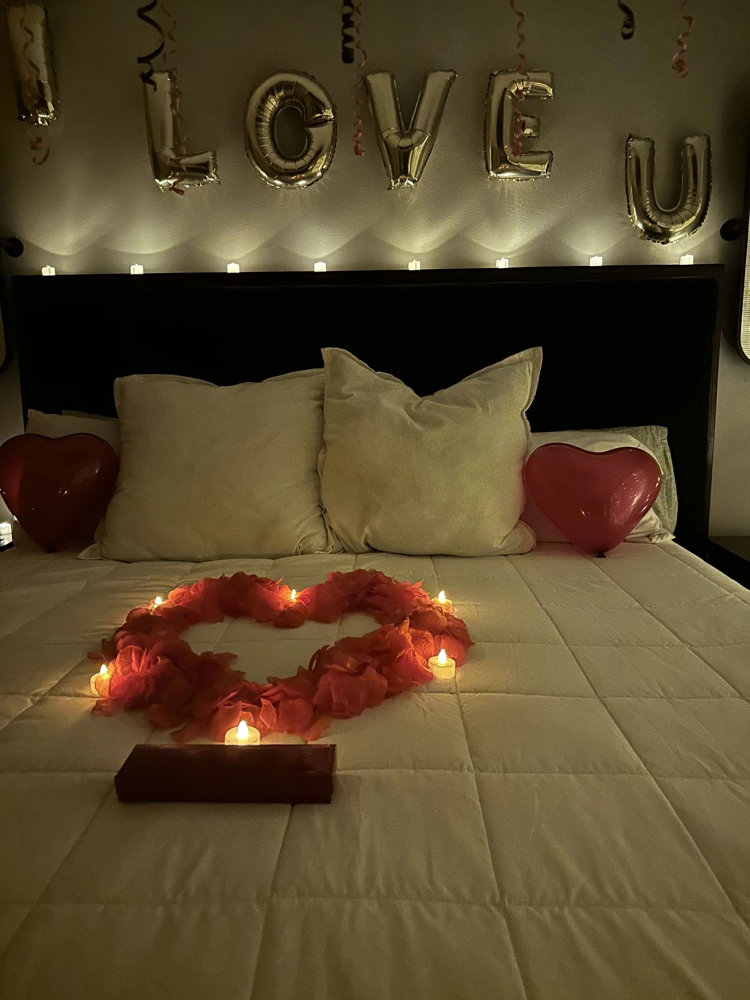 A decorated bedroom with a large bed that has white pillows and a white comforter. Above the bed, silver balloons spell out 'LOVE U,' and there are string lights. On the bed, there is a heart-shaped arrangement of pink rose petals surrounded by small