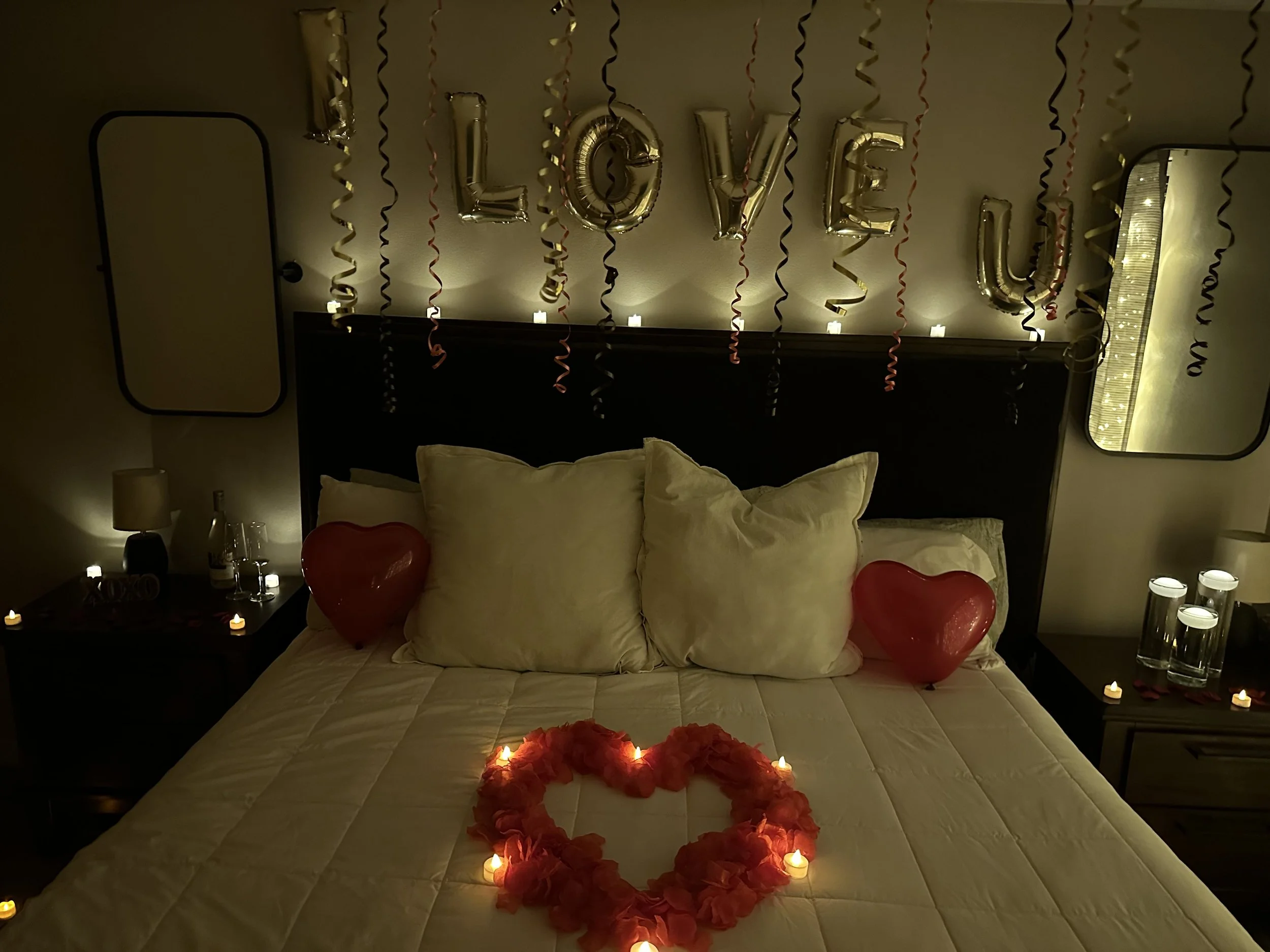 A decorated bedroom with love-themed decorations, including balloons, candles, and a heart shape made of flowers on the bed. The wall behind the bed has balloons spelling out 'LOVE YOU' and hanging streamers.
