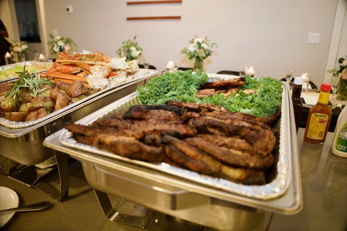 Buffet table with grilled meats, leafy greens, roasted potatoes, and crab legs at a party or gathering.