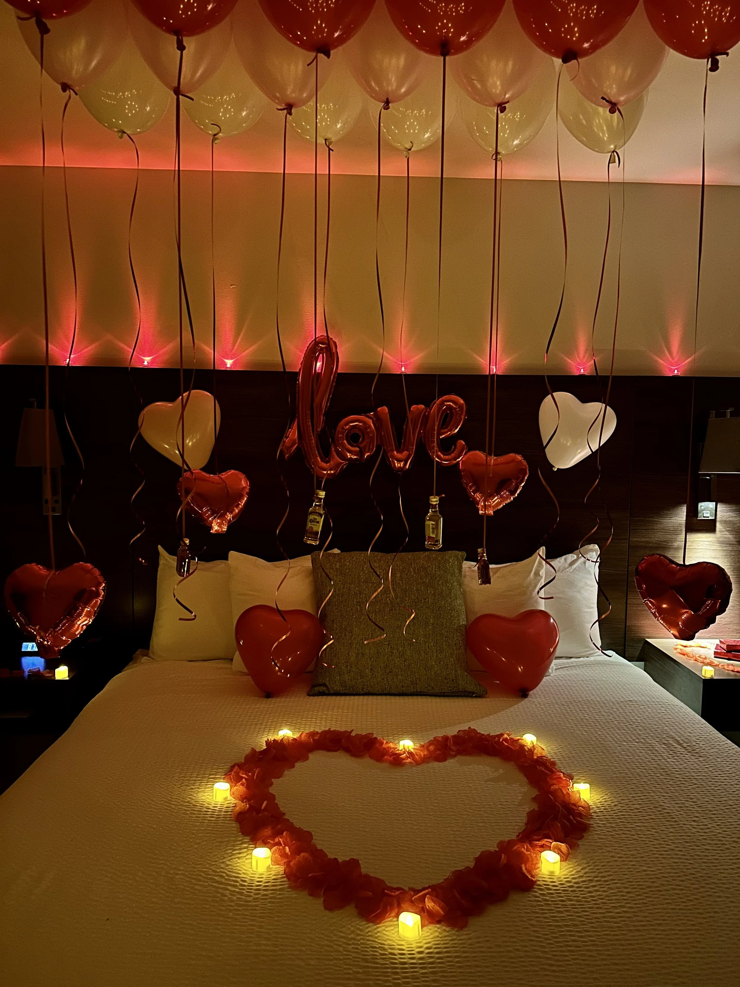 A romantic bedroom decorated with red and white balloons, heart-shaped balloons, candles, and an illuminated heart made of flower petals on the bed. The word 'love' is spelled out with balloons, and there are small bottles hanging from some balloons,