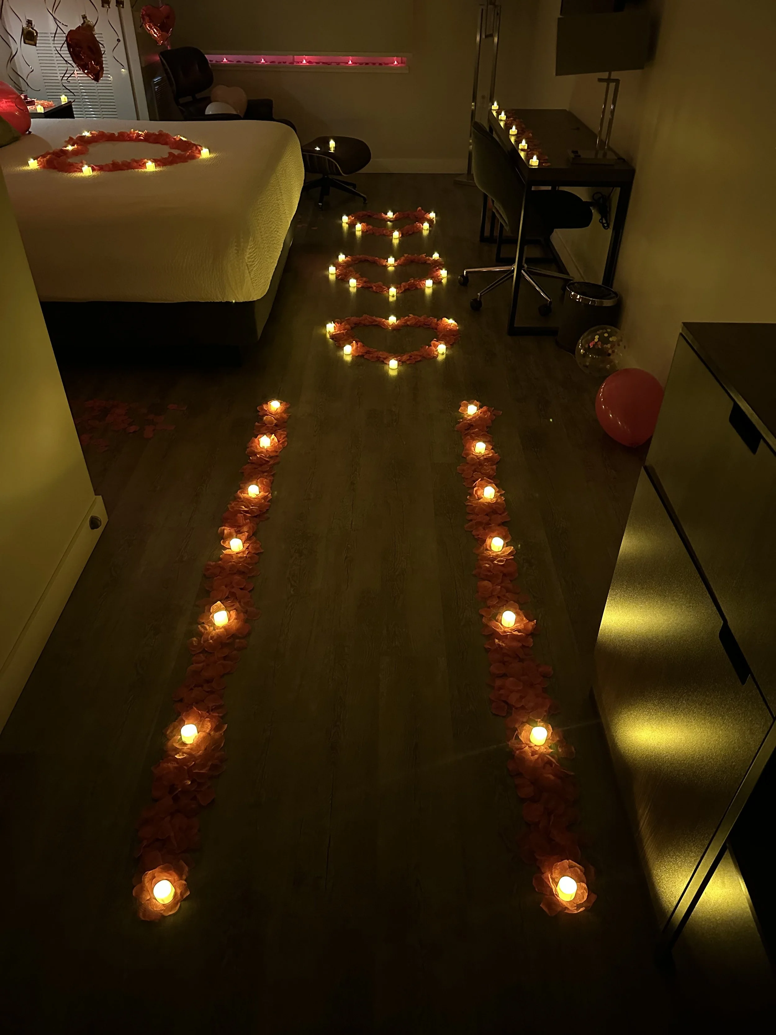 A dimly lit room decorated with candles and rose petals to form a path leading to a bed, suggesting a romantic setting.