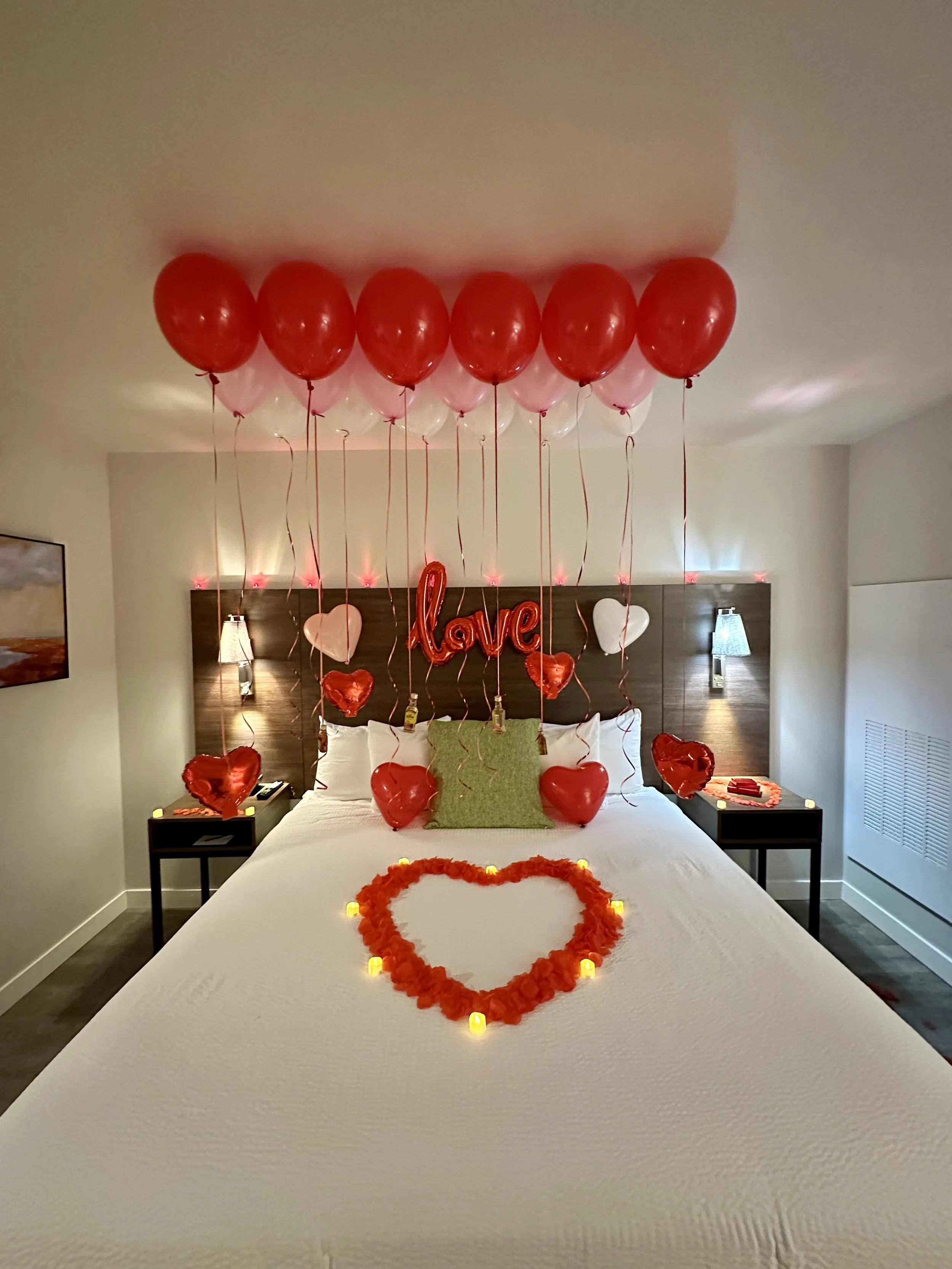 A hotel bed decorated for Valentine's Day with red and white balloons, heart-shaped balloons, a 'love' sign, candles, and a heart made of rose petals on the bed.