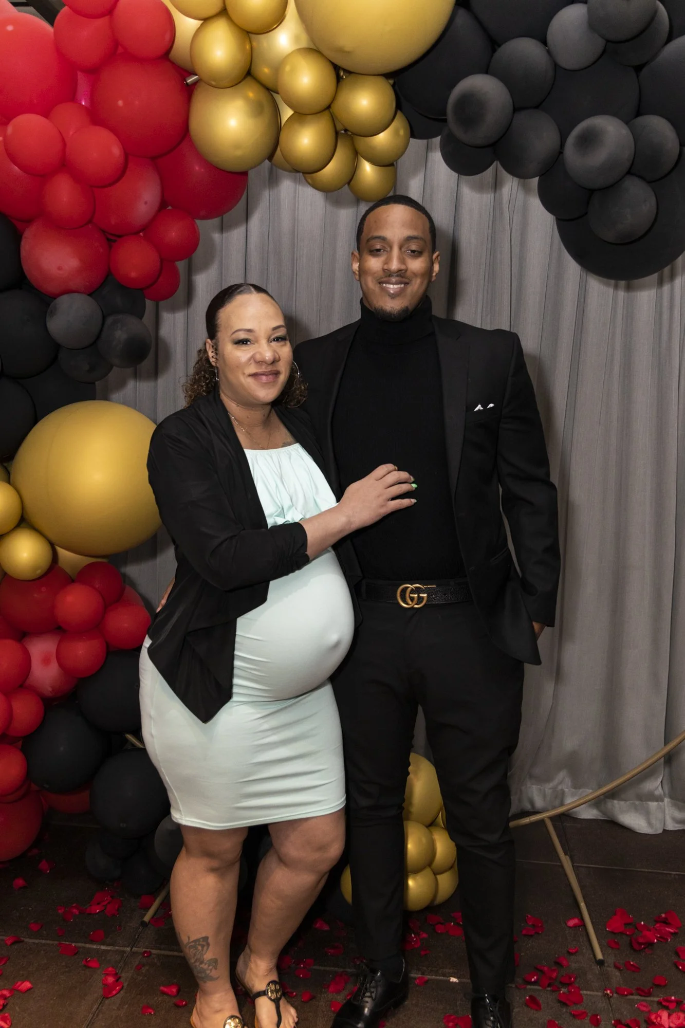 A pregnant woman and a man dressed in formal attire posing together at a celebration, with a backdrop of red, black, and gold balloons and rose petals on the floor.