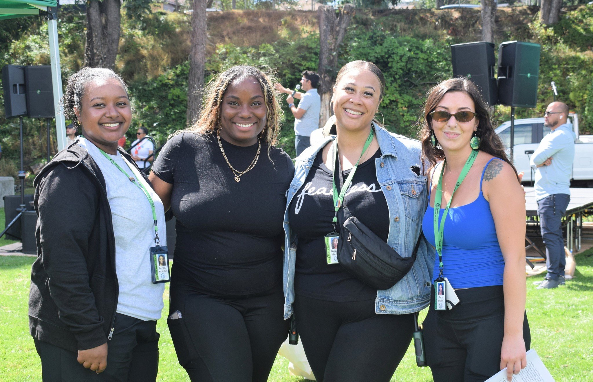 Four women standing outdoors smiling for the camera, with greenery and trees in the background, at a public event or gathering.