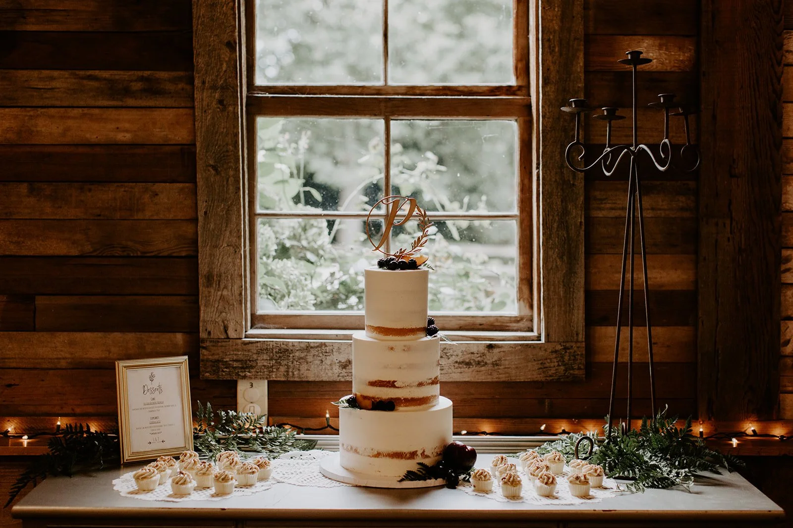Three-tiered wedding cake with semi-naked frosting, decorated with berries and a monogram topper, surrounded by cupcakes, greenery, and a framed menu, set against a wooden wall and window.