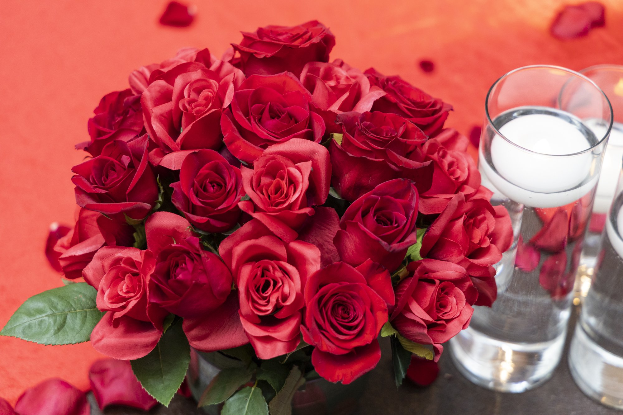 Bouquet of red roses with green leaves and several tall glasses of water with floating rose petals.