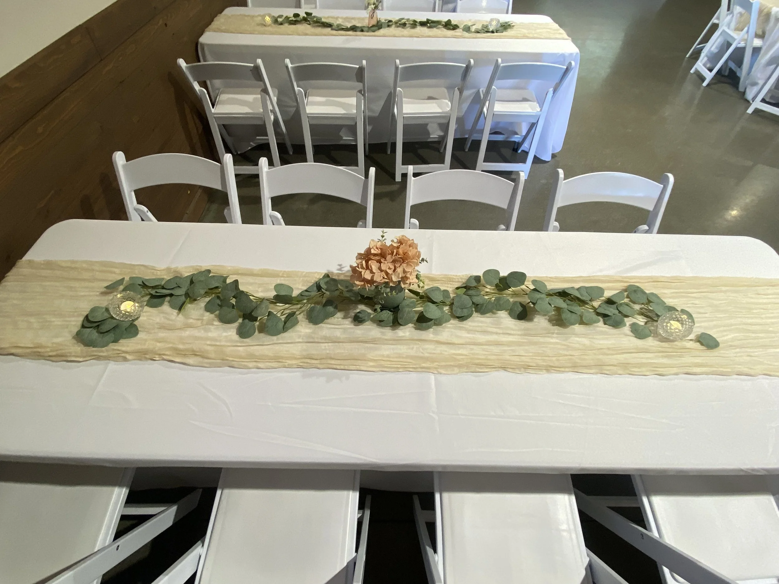 Event table decorated with a beige runner, eucalyptus leaves, a peach-colored flower centerpiece, and glass candle holders, surrounded by white chairs.