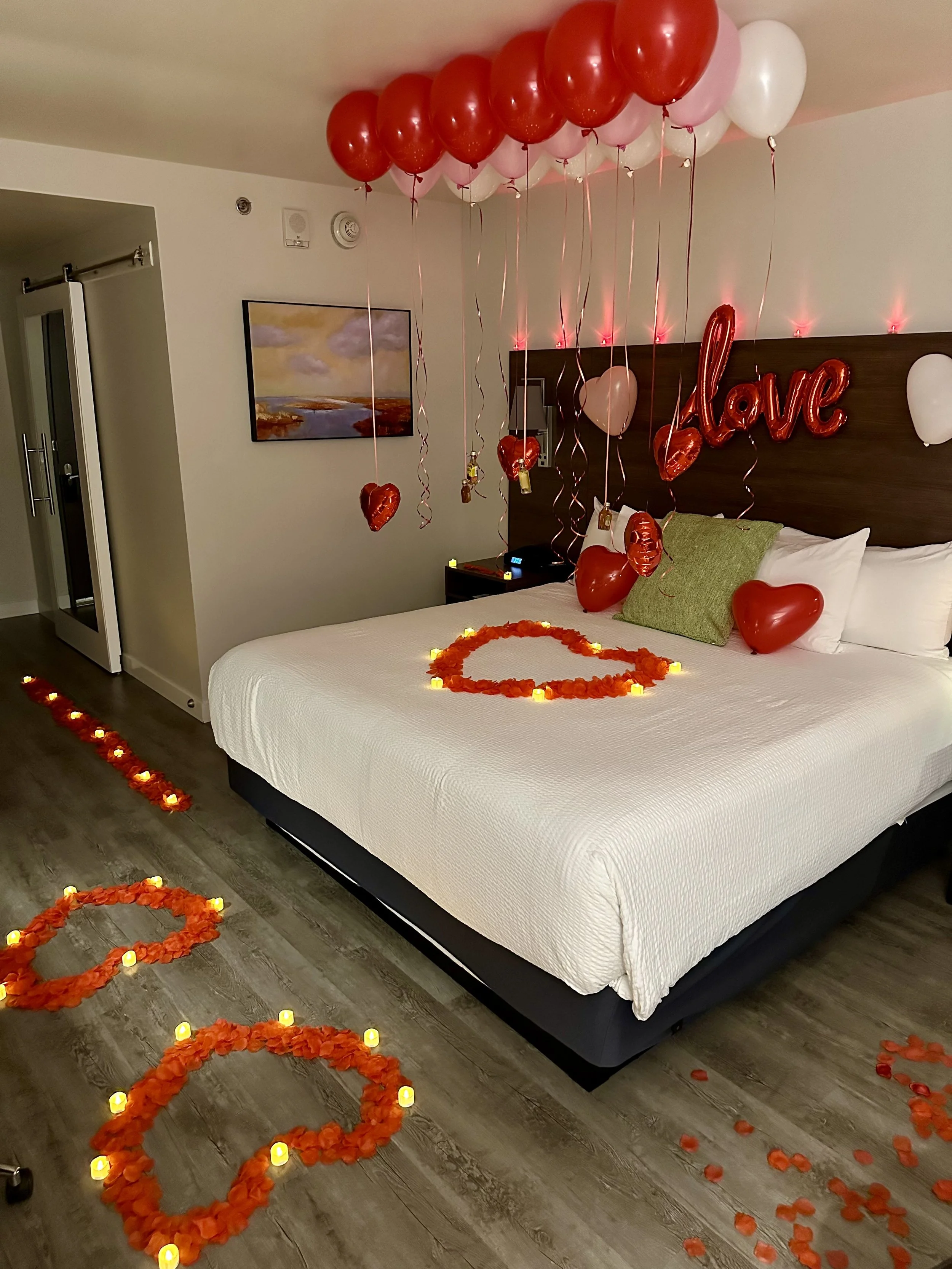 Romantic hotel room decorated with red and white balloons, heart-shaped balloons, rose petals, candles, and a love sign for a celebration of love.