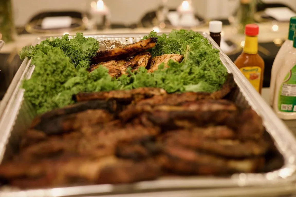 A tray of grilled meats, including ribs and chicken, garnished with fresh curly lettuce leaves, in a kitchen or dining setting.