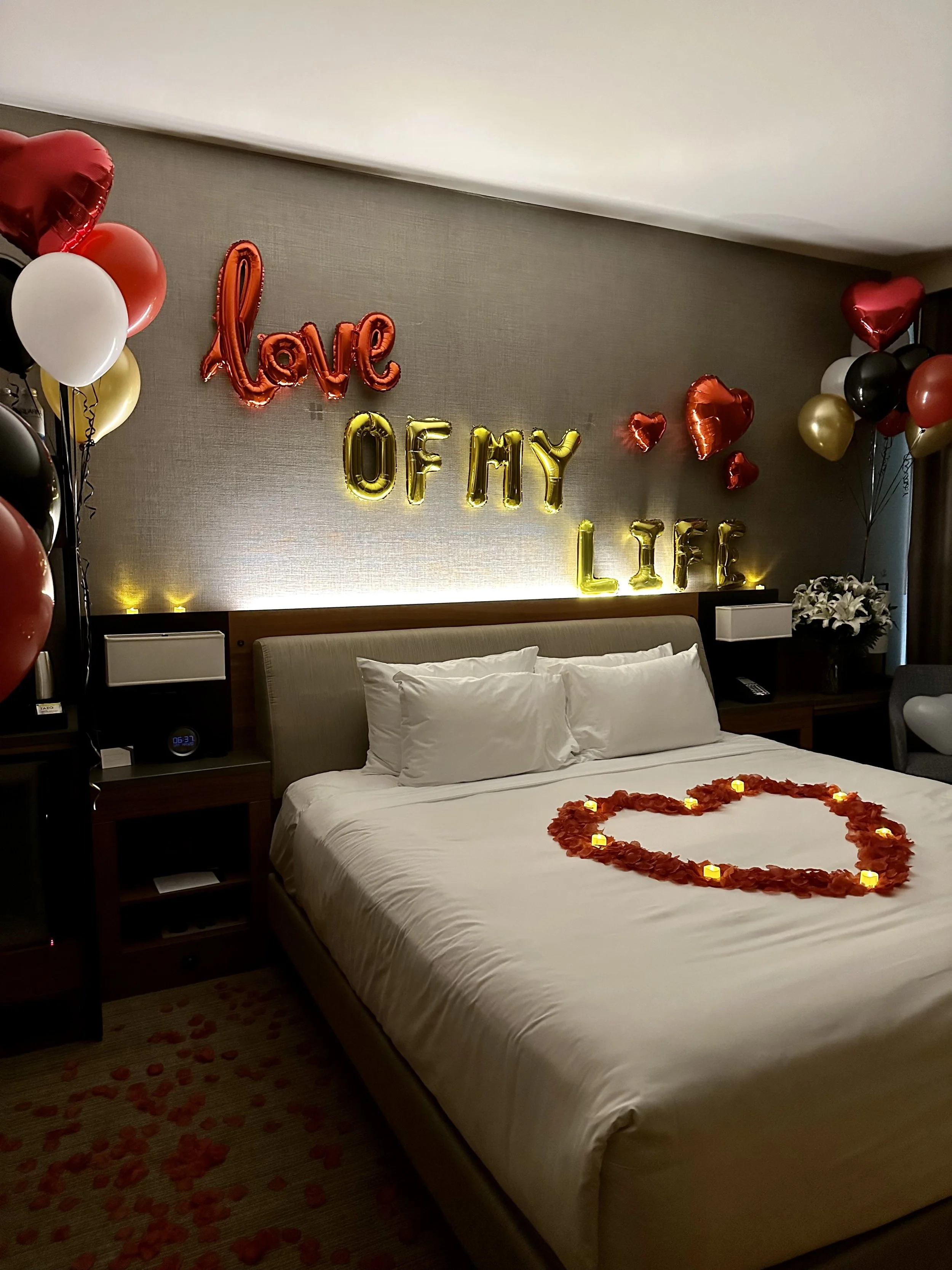 Hotel room decorated with heart-shaped balloons, string lights spelling "love" and "life," and a heart-shaped arrangement of rose petals and candles on the bed, celebrating love and life.