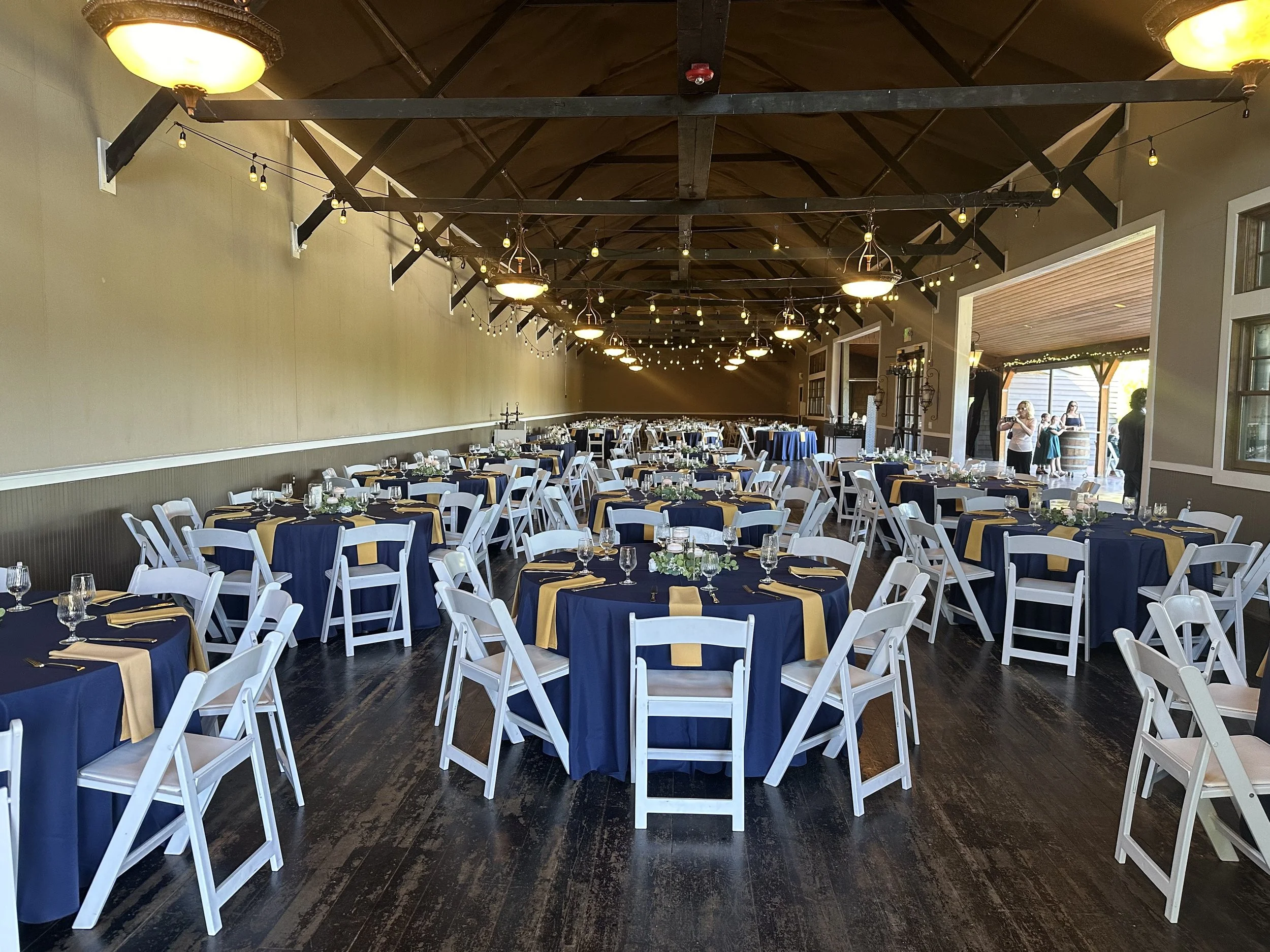 Wedding reception hall with round tables covered with navy blue tablecloths and yellow accents, white chairs, and elegant table settings, decorated with string lights hanging from the ceiling.