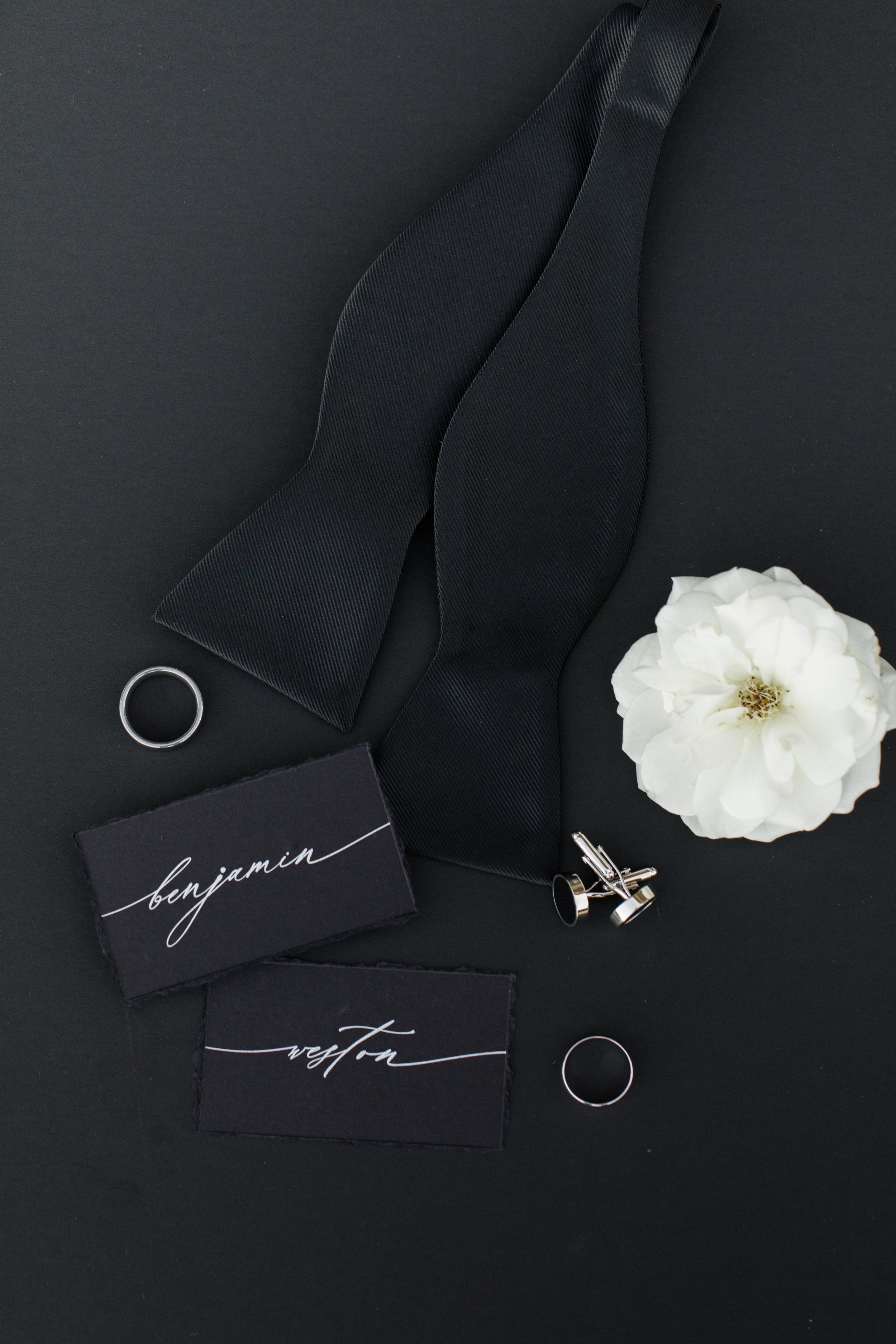 Wedding accessories including wedding rings, cufflinks, black bow tie, and black name cards with white calligraphy, all arranged on a black background with a white flower.