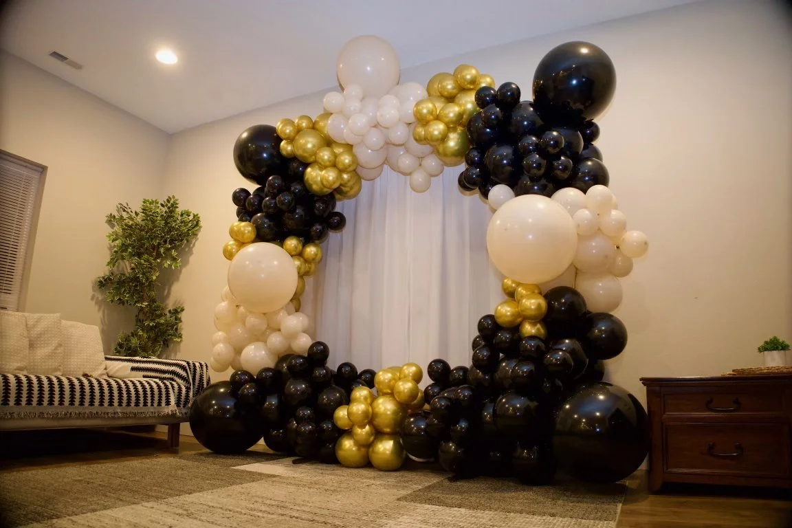 Balloon decoration in black, white, and gold surrounding a white curtain in a living room.