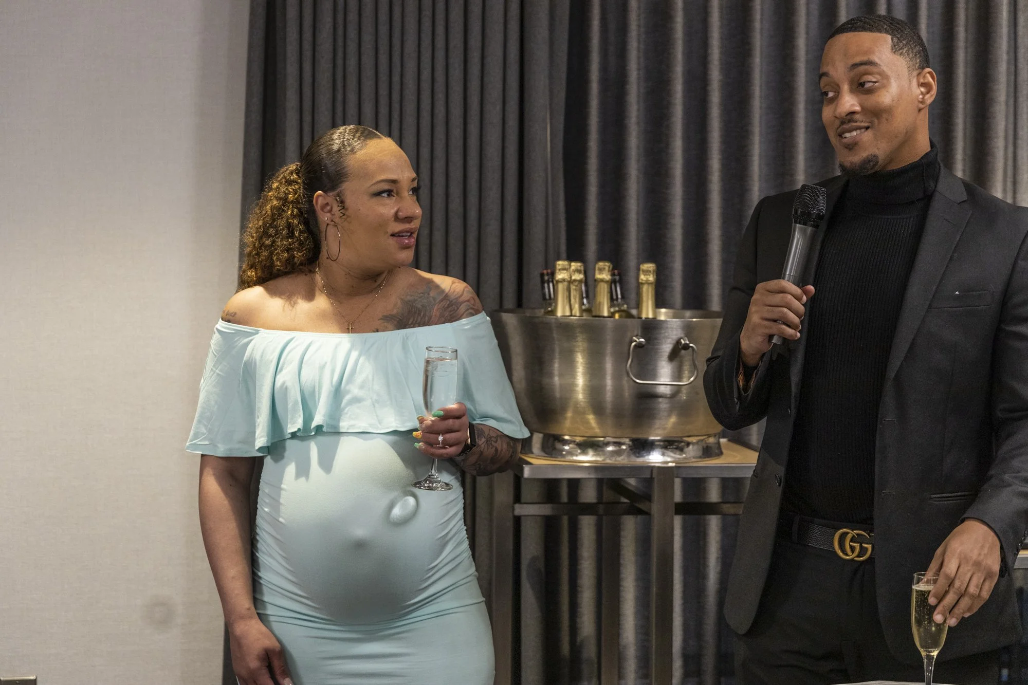 A woman in a light blue off-the-shoulder dress holding a champagne glass, looking at a man in a black suit with a turtleneck who is speaking into a microphone at a social gathering.