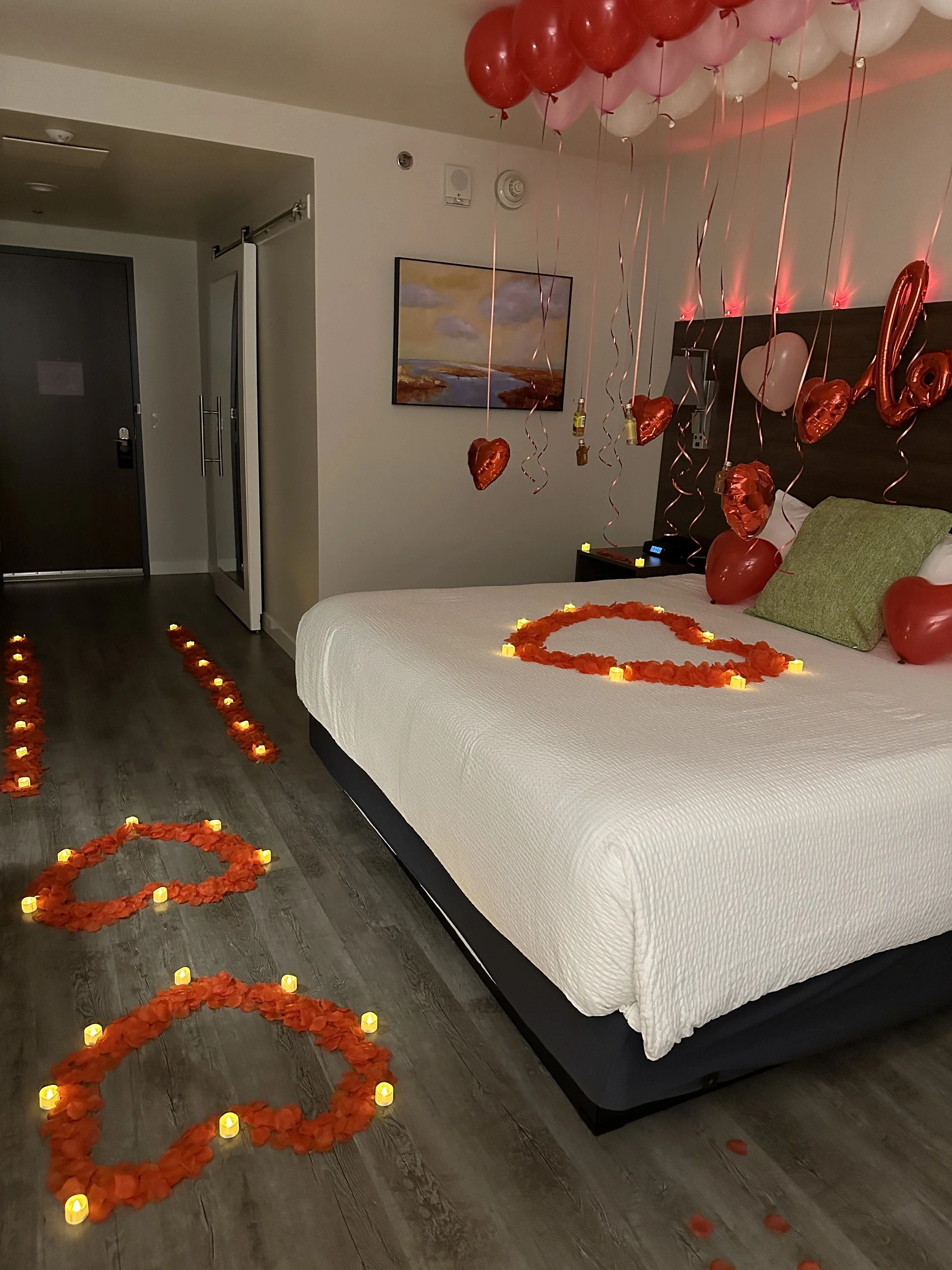 Hotel room decorated for Valentine's Day with heart-shaped flower and candle arrangements on the bed and floor, and red and pink balloons hanging from the ceiling.