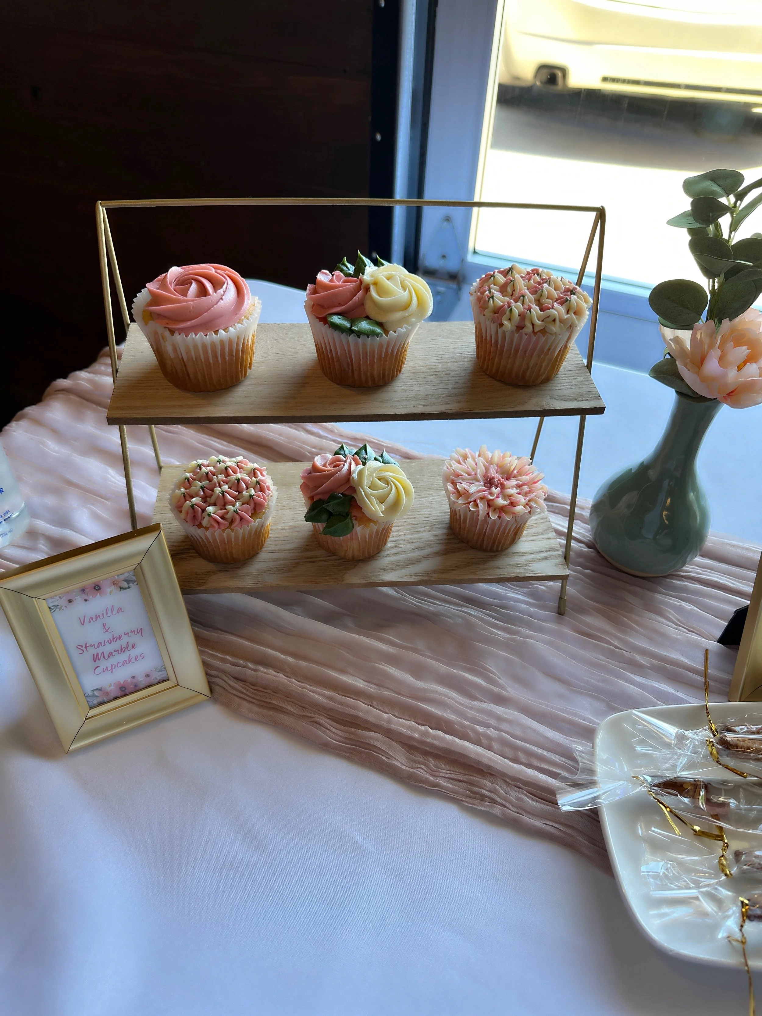 Display of vanilla and strawberry marble cupcakes with pink and white frosting floral decorations on a two-tiered wooden and gold stand, on a table with a pink fabric runner, a framed sign, and a white plate with wrapped treats, near a window with a 