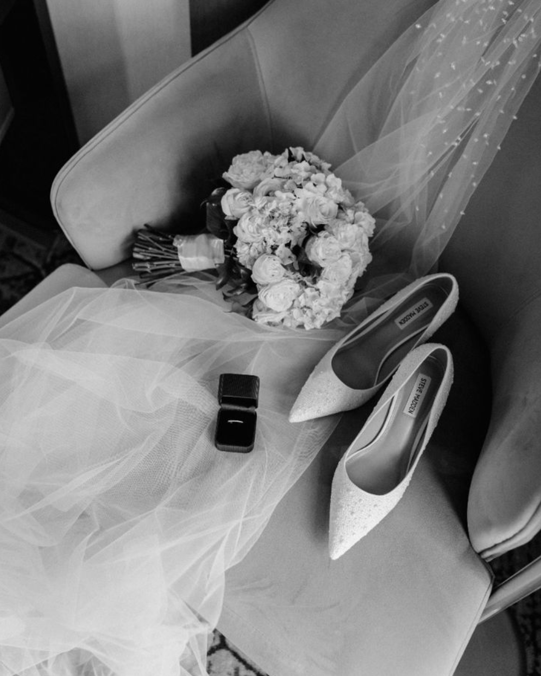 A wedding bouquet, a pair of white high heels, a small black box, and a sheer veil are arranged on a chair, likely for a bride.