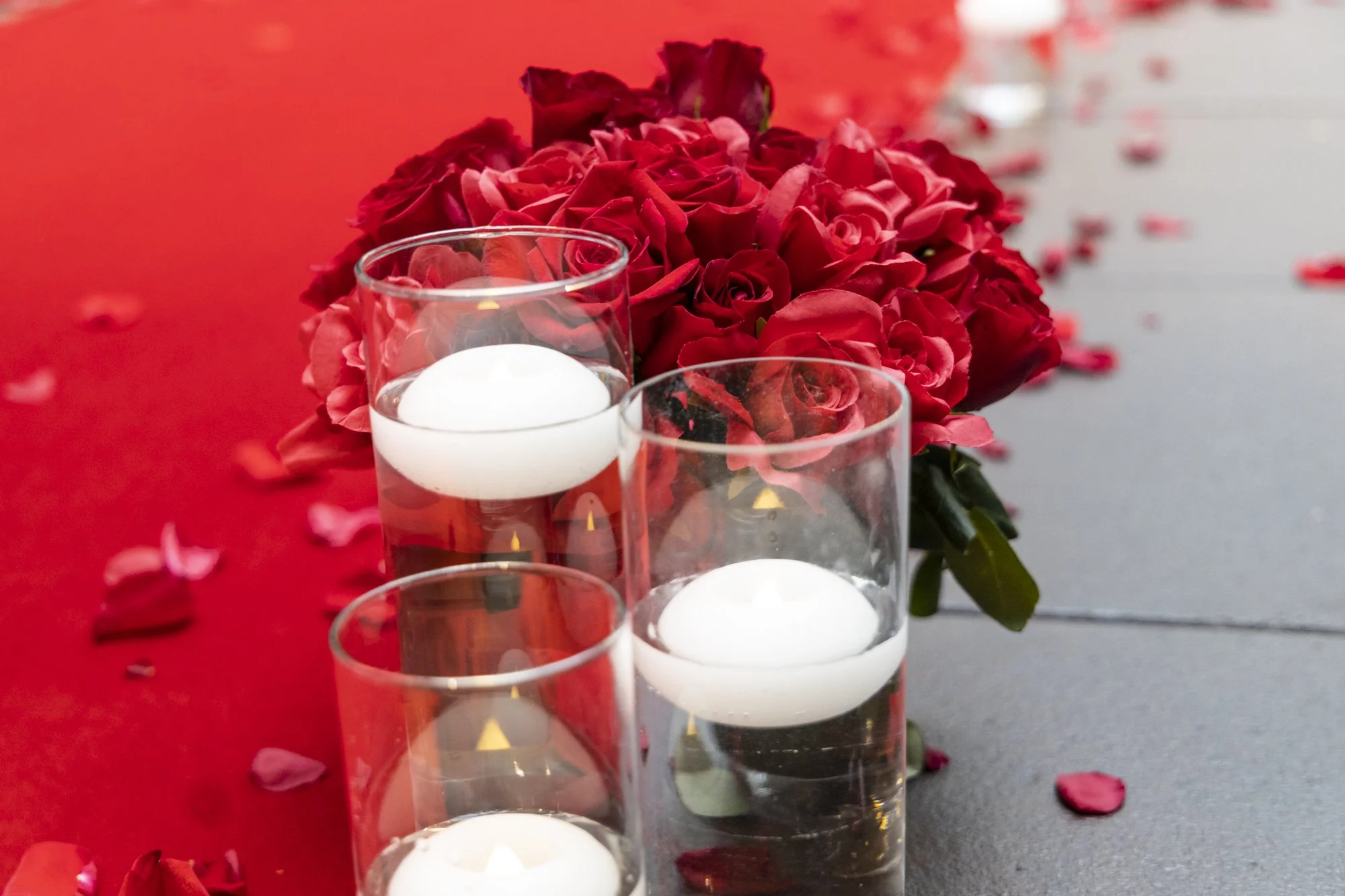 Red roses and white floating candles on a table with rose petals scattered around, possibly for a romantic or memorial setting.
