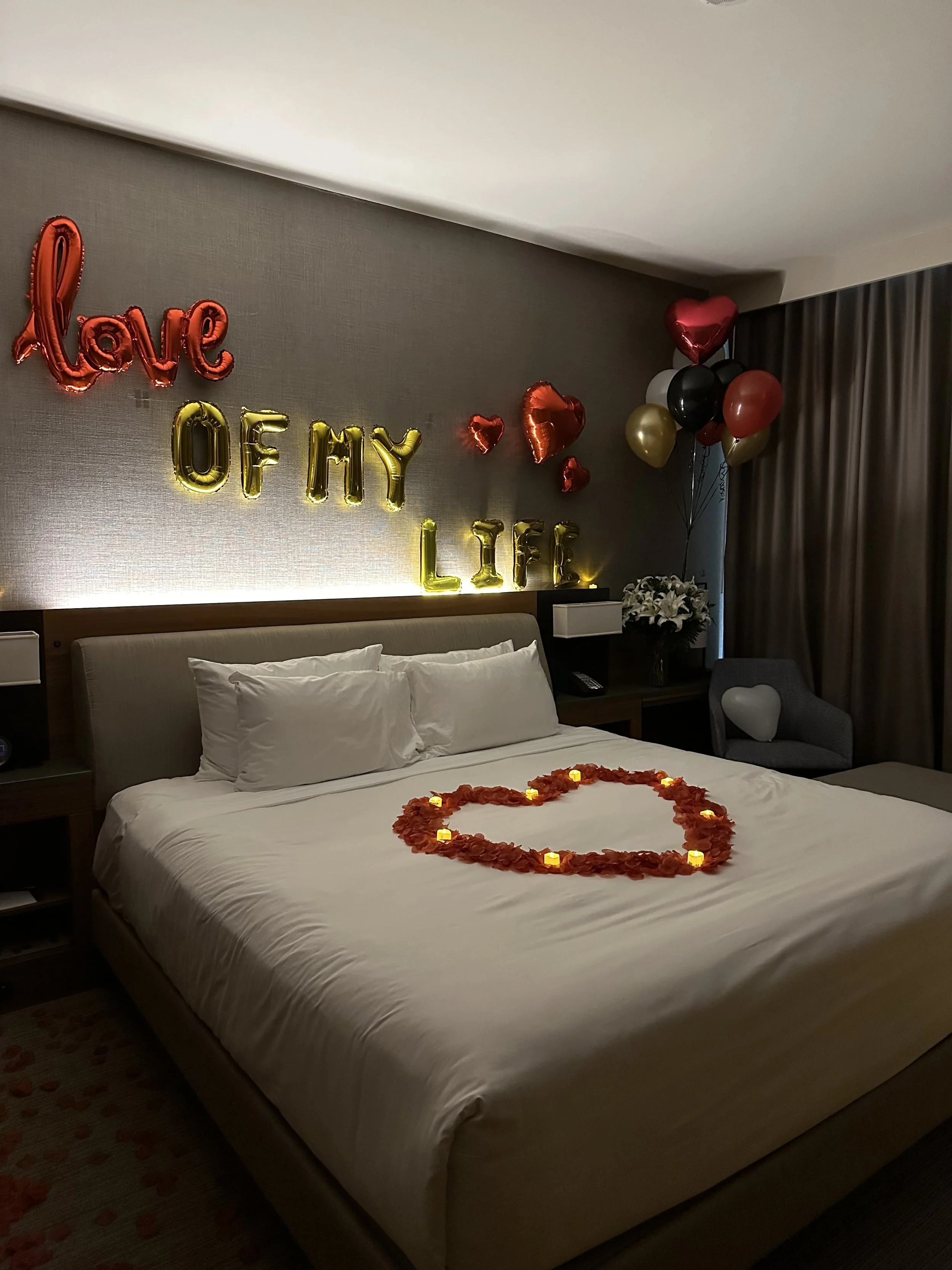 Decorated hotel room with balloons spelling 'love of my life' on the wall, heart-shaped balloons, a large heart made of rose petals and small candles on the bed, and a chair with a heart-shaped pillow.