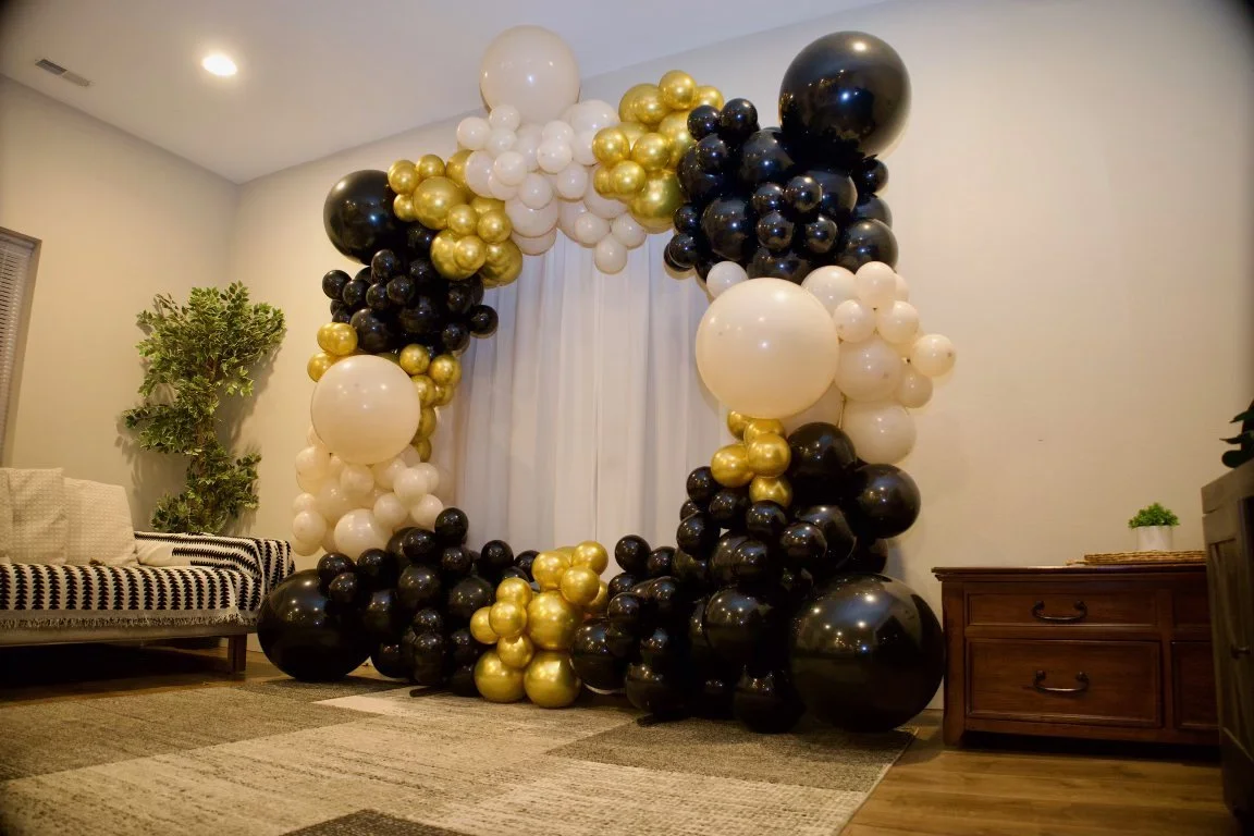 Black, white, and gold balloons arranged in a large arch in a living room with a sofa, indoor plants, a curtain, and a wooden cabinet.