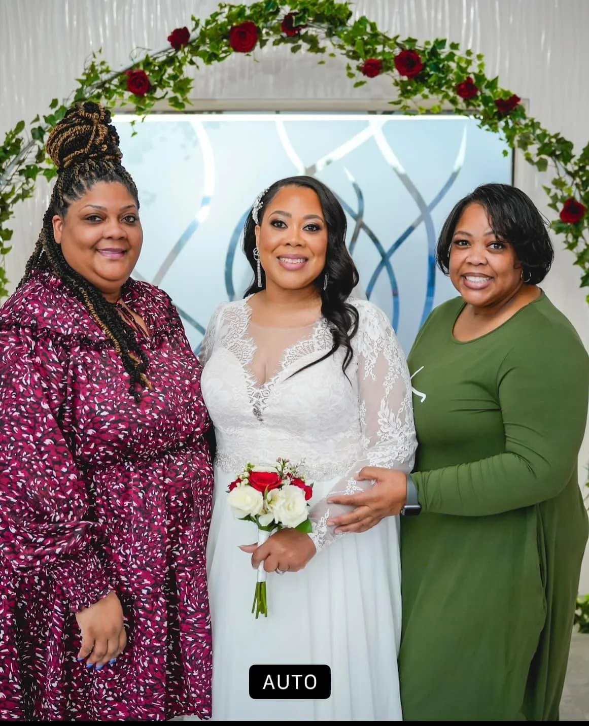A bride in a white lace wedding dress holding a bouquet of red and white roses, flanked by two women, one in a purple patterned dress and the other in a green dress, standing under a floral arch with red roses.