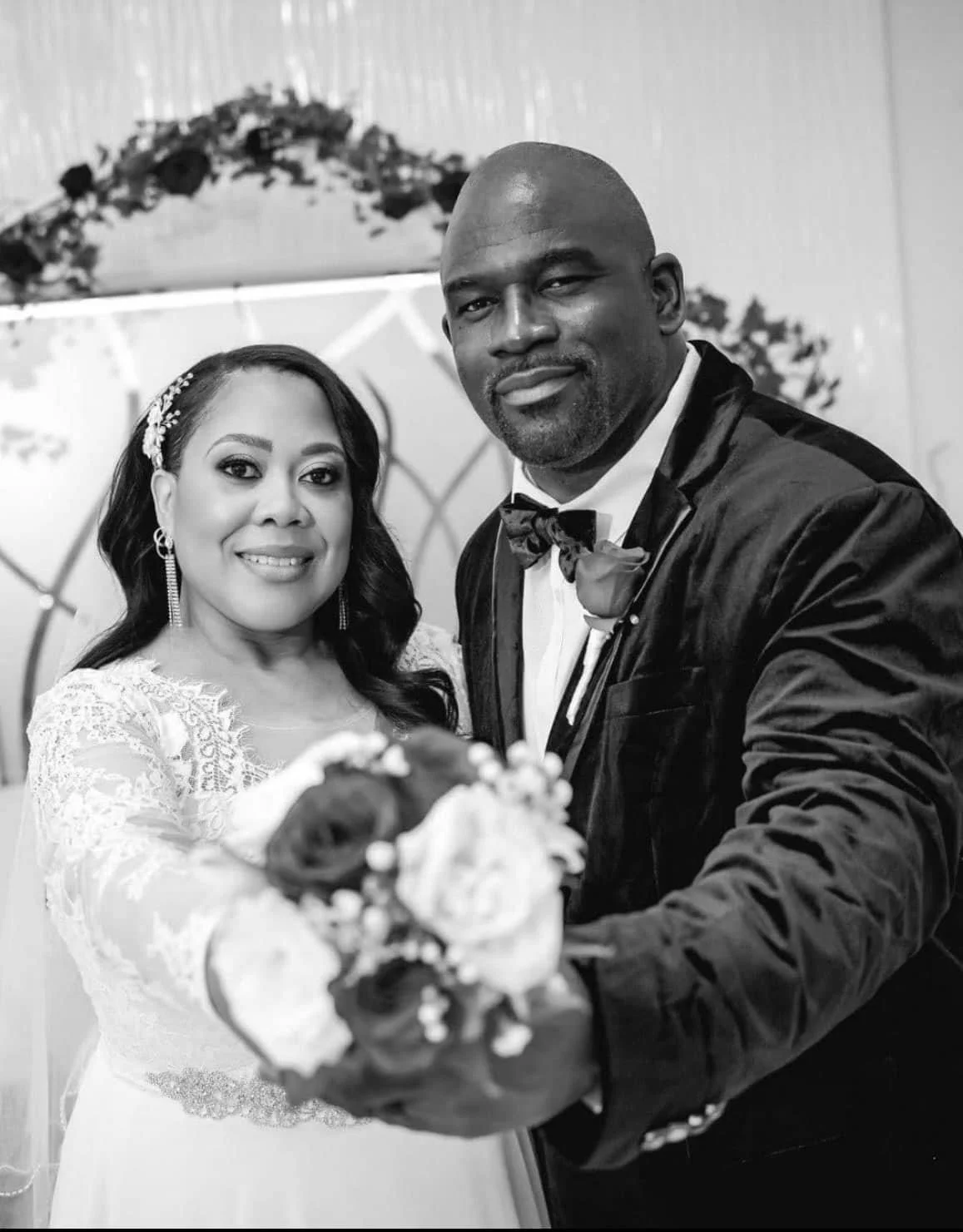 Black and white photo of a bride and groom on their wedding day, holding a bouquet of flowers, with the bride smiling and the groom looking at the camera.