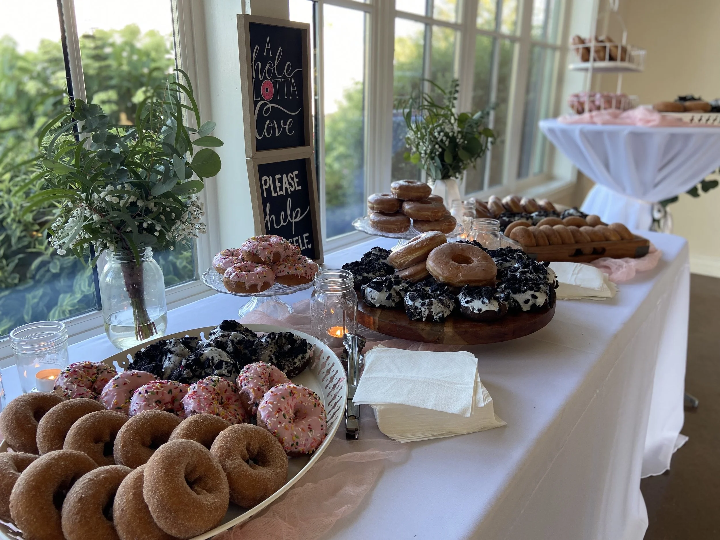 A table with assorted donuts and cookies at a party or event, decorated with flowers and signs that read 'A hole lotta love' and 'Please help yourself'.