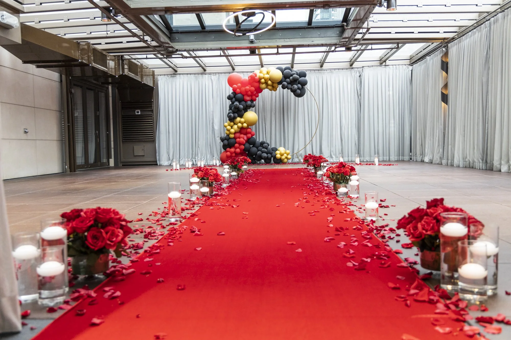 Indoor wedding aisle decorated with red rose petals, candles, and floral arrangements leading to a balloon backdrop with a circular frame.