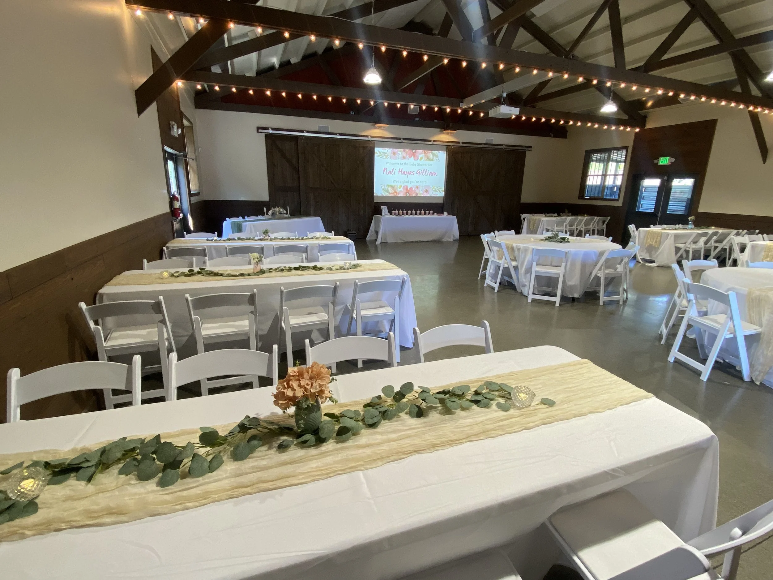 Event hall decorated with white tablecloths, eucalyptus garlands, and floral centerpieces, set up for a celebration or reception, with a projection screen at the front displaying the name 'Nali Hayes William'.