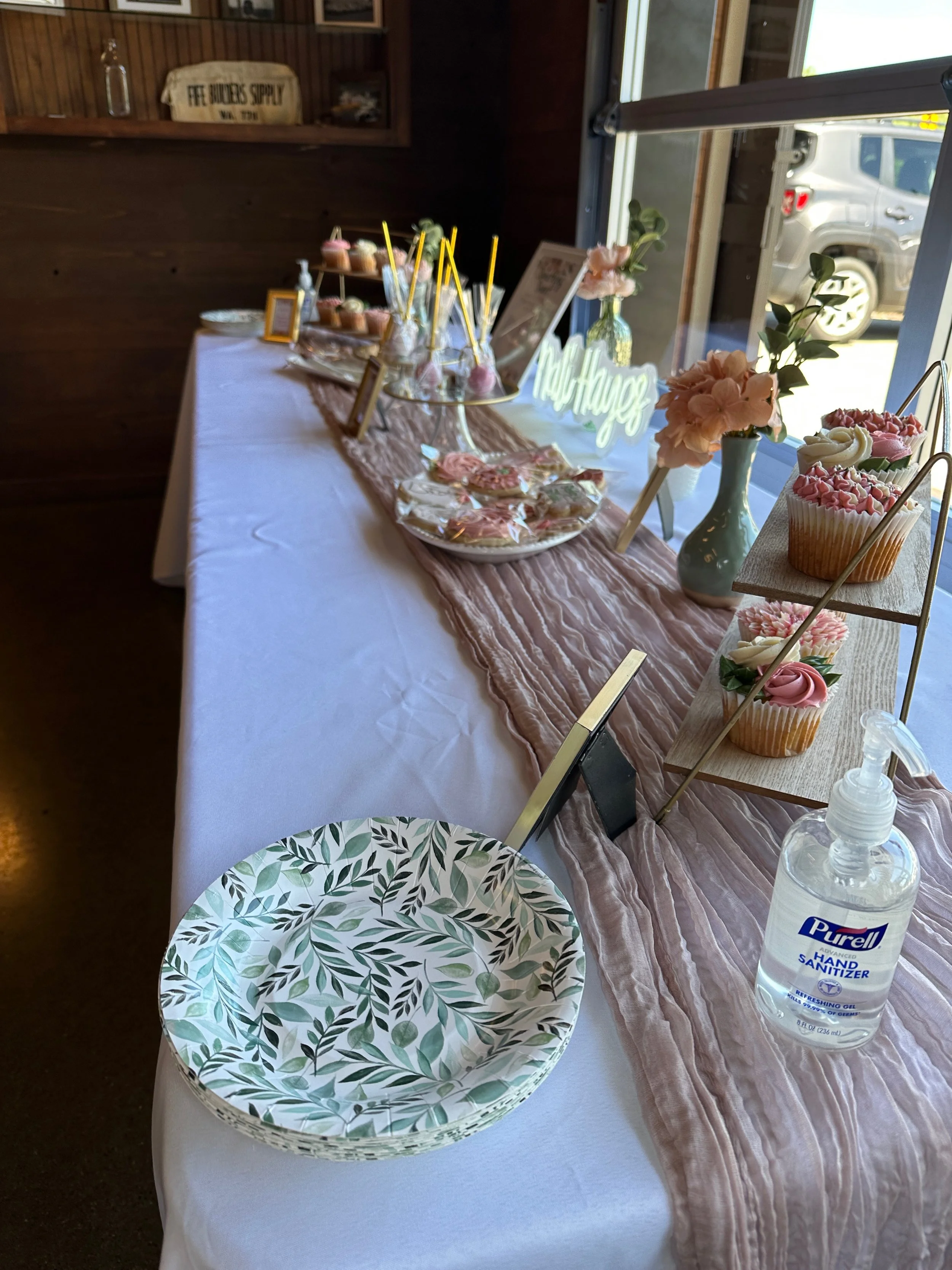 Sweet table decorated with pink and white cupcakes, lollipops, flower arrangements, a hand sanitizer bottle, and a pink table runner.
