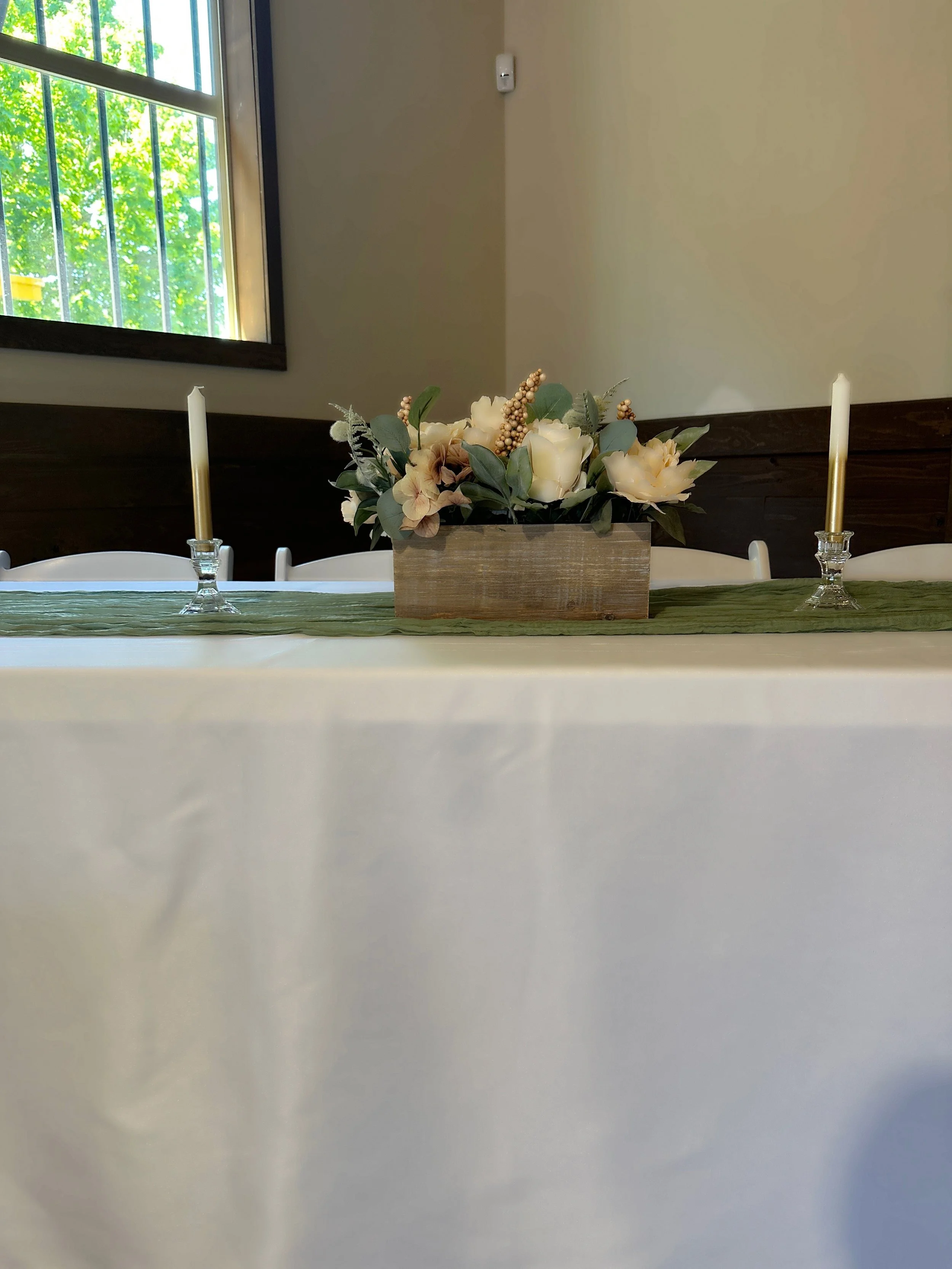 Centered floral centerpiece with white roses and greenery on a table with a green table runner, flanked by two white candles in glass holders, in a room with a window and beige and dark wood walls.
