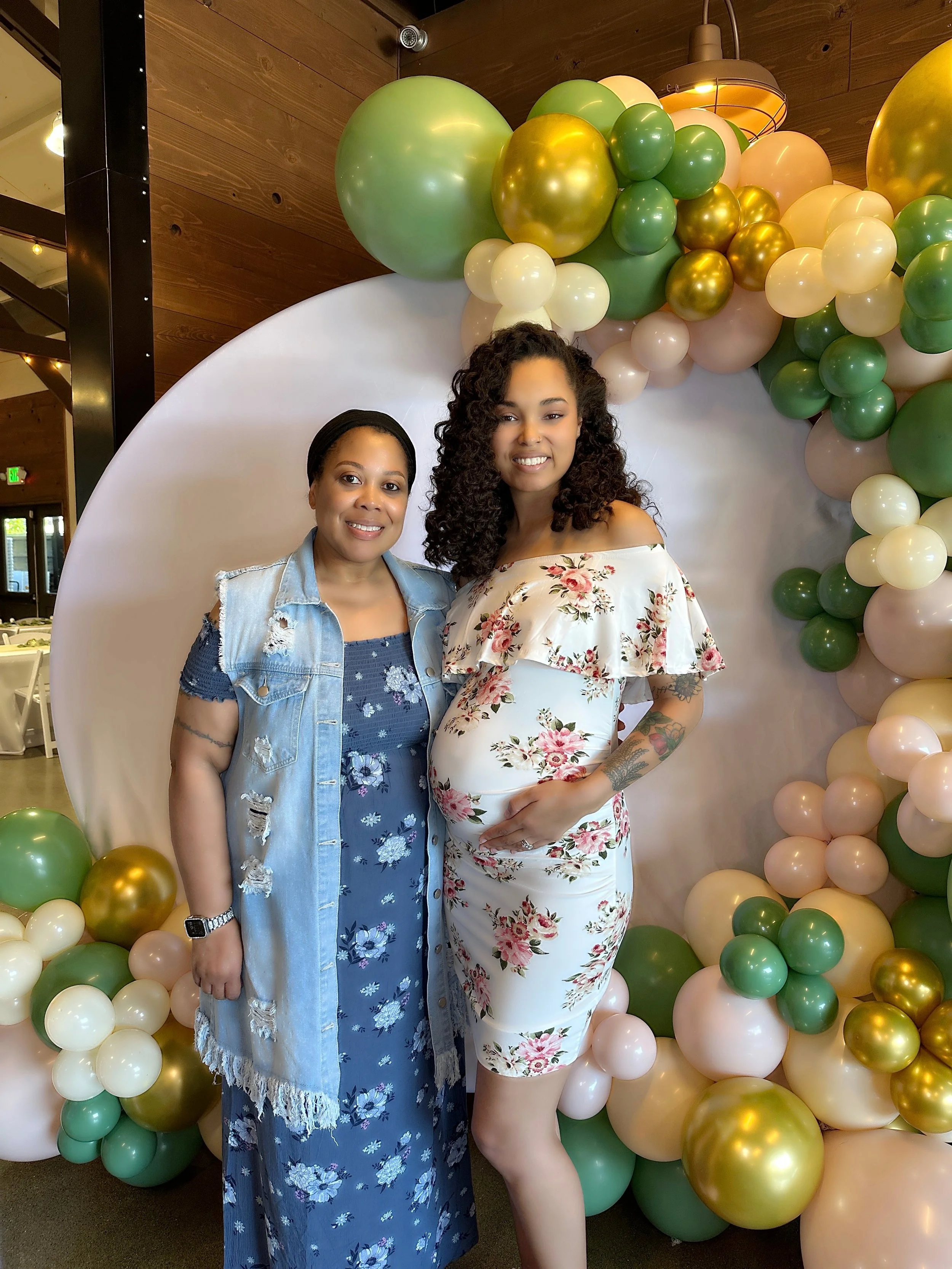 A woman and a pregnant woman smiling and standing together in front of a balloon decoration. The woman on the left is wearing a blue floral dress with a distressed denim vest, and the pregnant woman on the right is wearing an off-shoulder floral dres