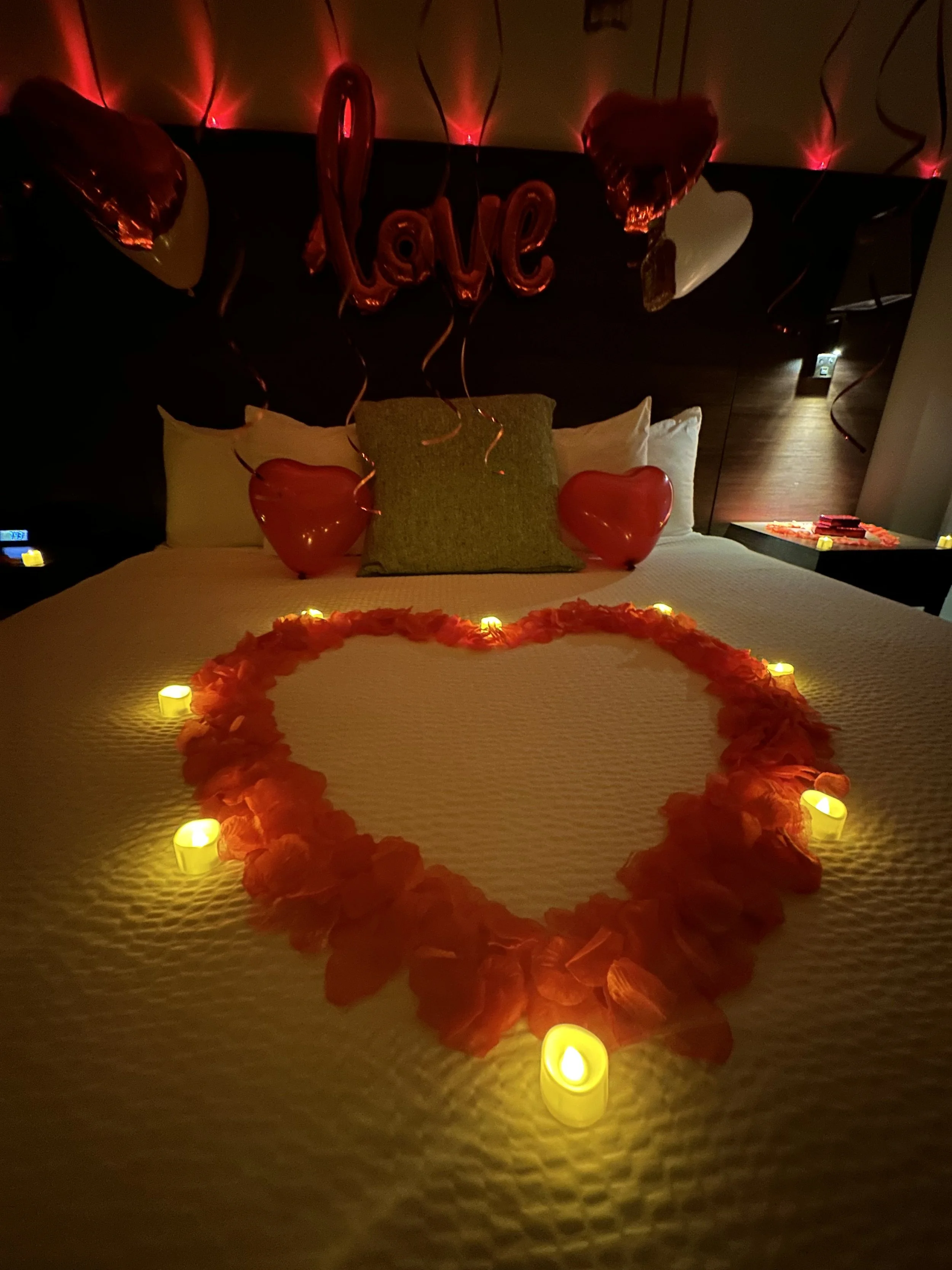 A romantic bedroom decorated with red and yellow balloons, candles, and a heart-shaped flower arrangement on the bed. Latex balloons spell out "love" hanging from the ceiling, creating a cozy atmosphere.