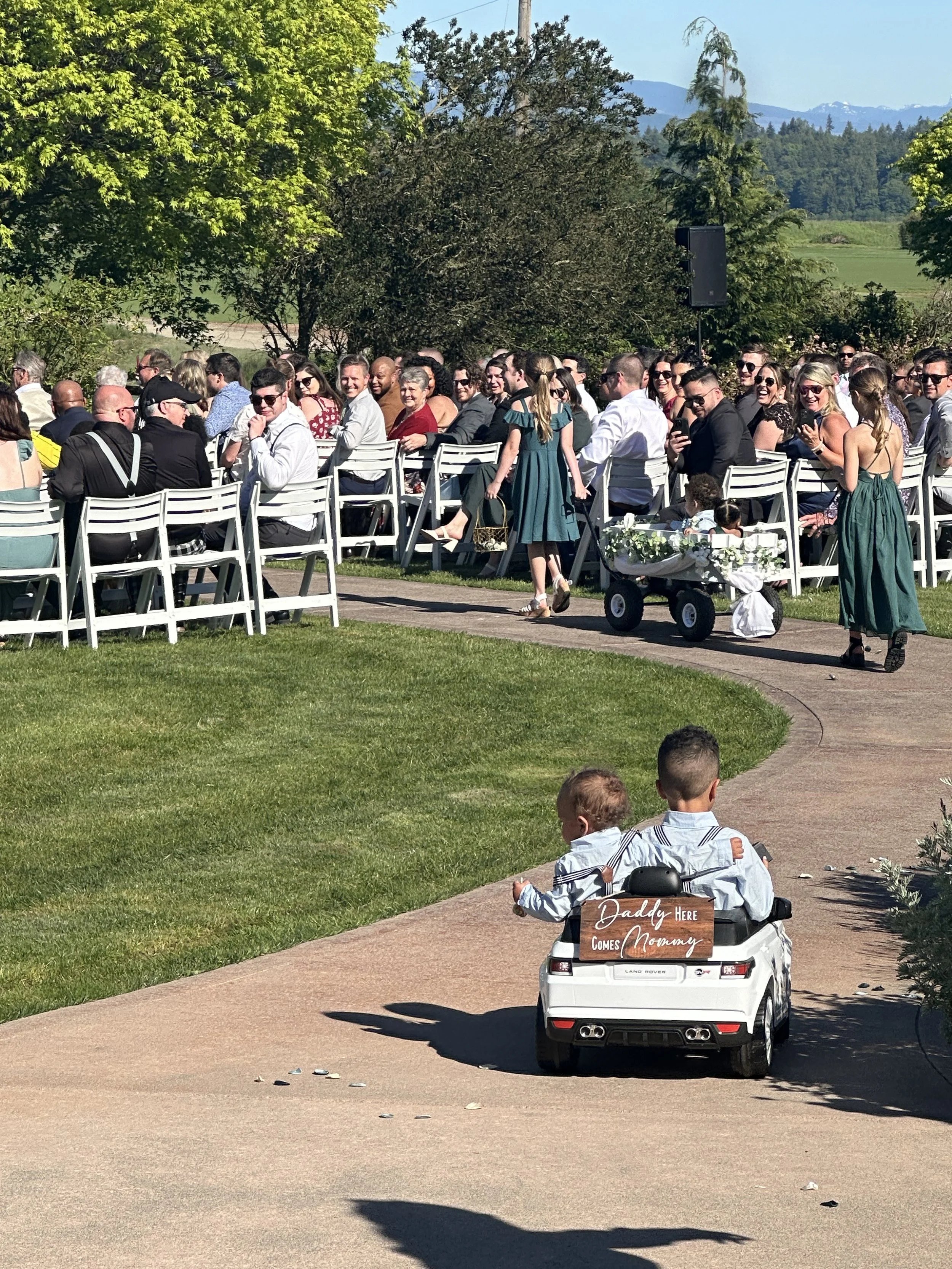 Children riding a toy car with a sign saying "Daddy Here Comes Mommy," at an outdoor wedding reception with many guests seated in white chairs, surrounded by green trees and mountains in the background.