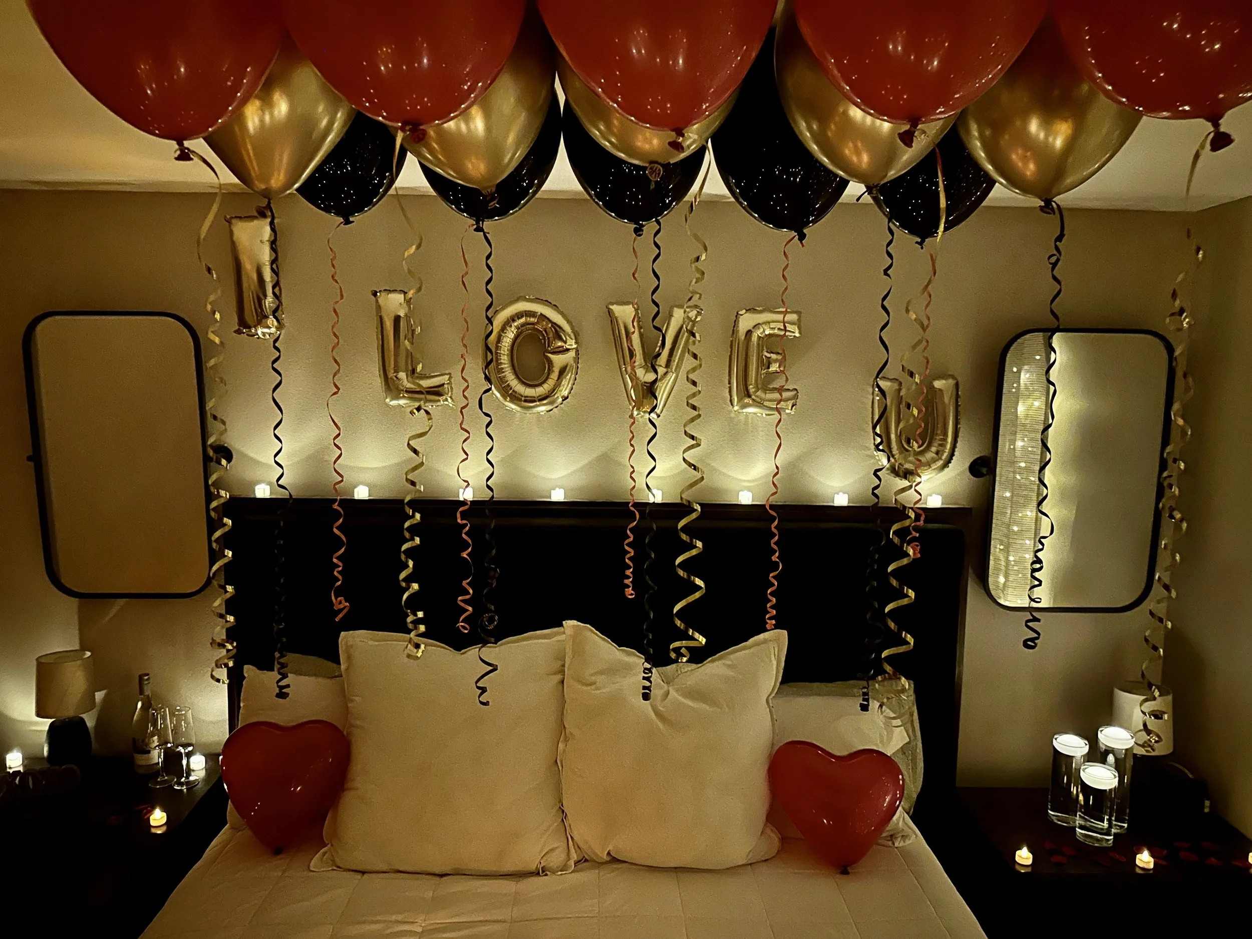 Decorated bedroom with red and black balloons, gold 'LOVE' balloons, and red heart-shaped balloons on the bed, indicating a romantic celebration.