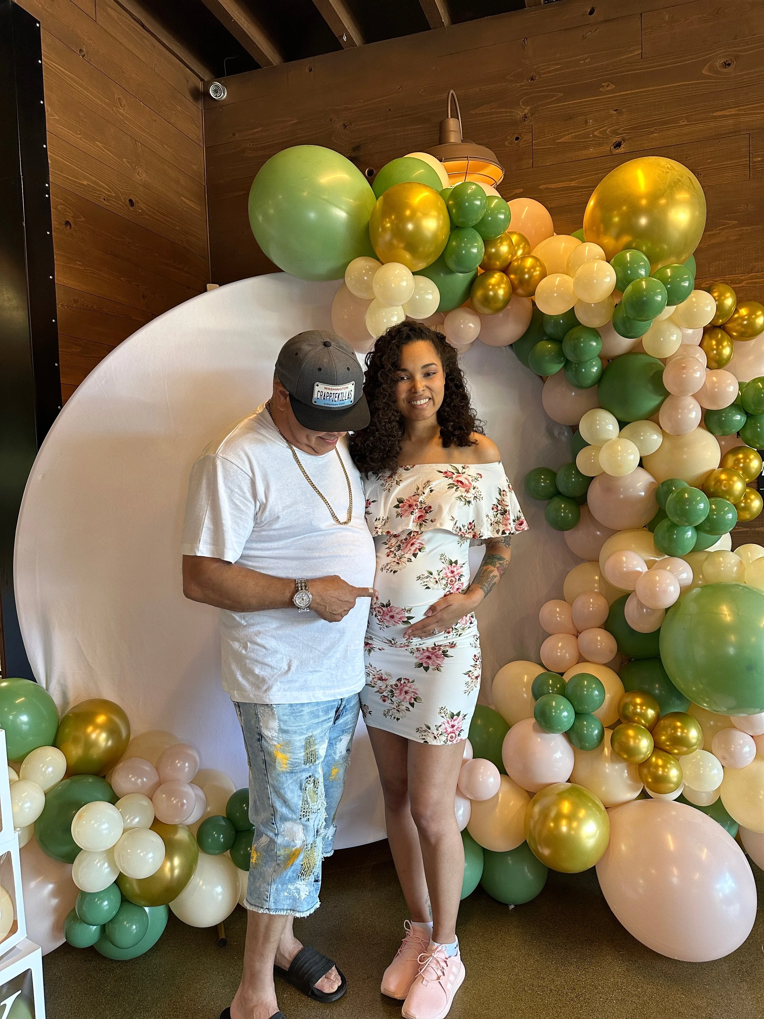 A pregnant woman with curly hair in a floral dress standing next to a man in a white t-shirt, ripped jeans, and a black cap. They are posing in front of a balloon arch with green, gold, and pastel-colored balloons. The woman is smiling and resting he