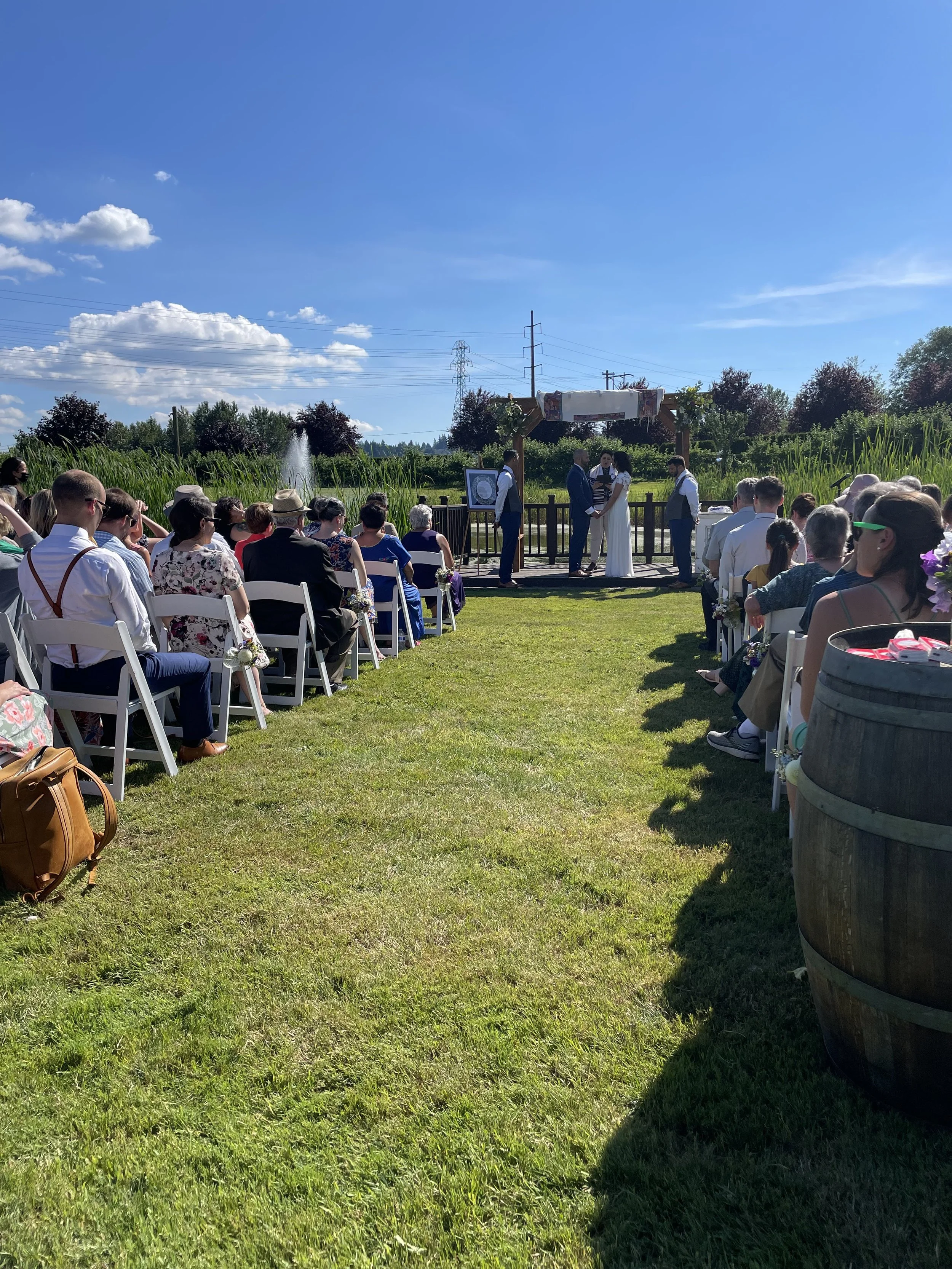 Outdoor wedding ceremony with guests seated on white chairs facing an altar, under a bright blue sky with scattered clouds, surrounded by greenery and tall grasses.