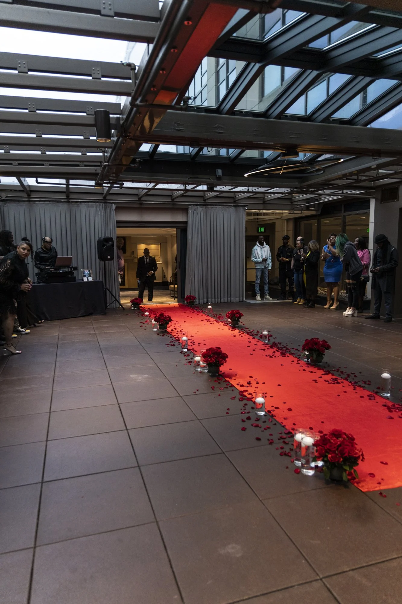 Indoor wedding ceremony setup with a red carpet, flower arrangements, and candle lanterns on a tiled floor, with guests gathered around.