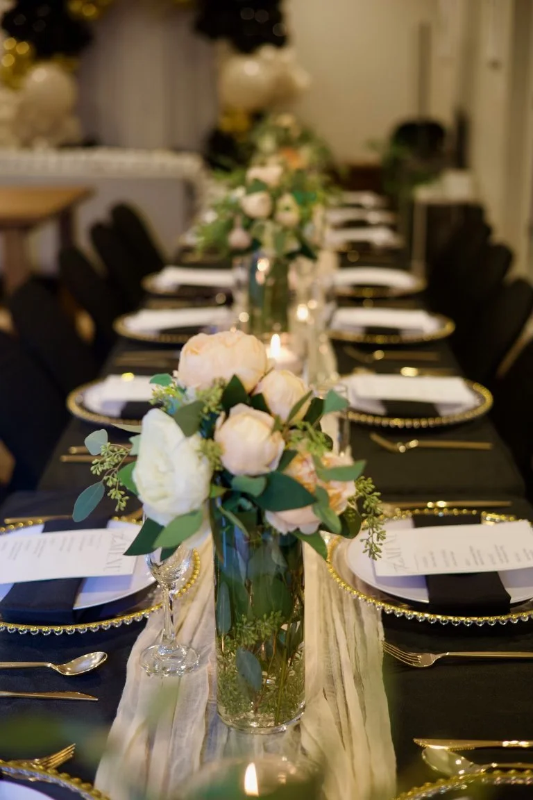 Elegant banquet table decorated with floral centerpieces, candles, black and gold plates, gold utensils, and black napkins, set for a formal event.